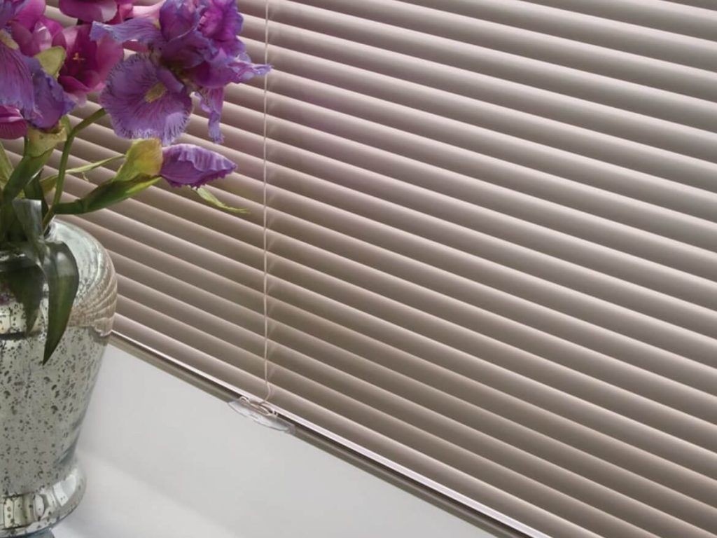A vase of purple flowers is sitting in front of a window with blinds.
