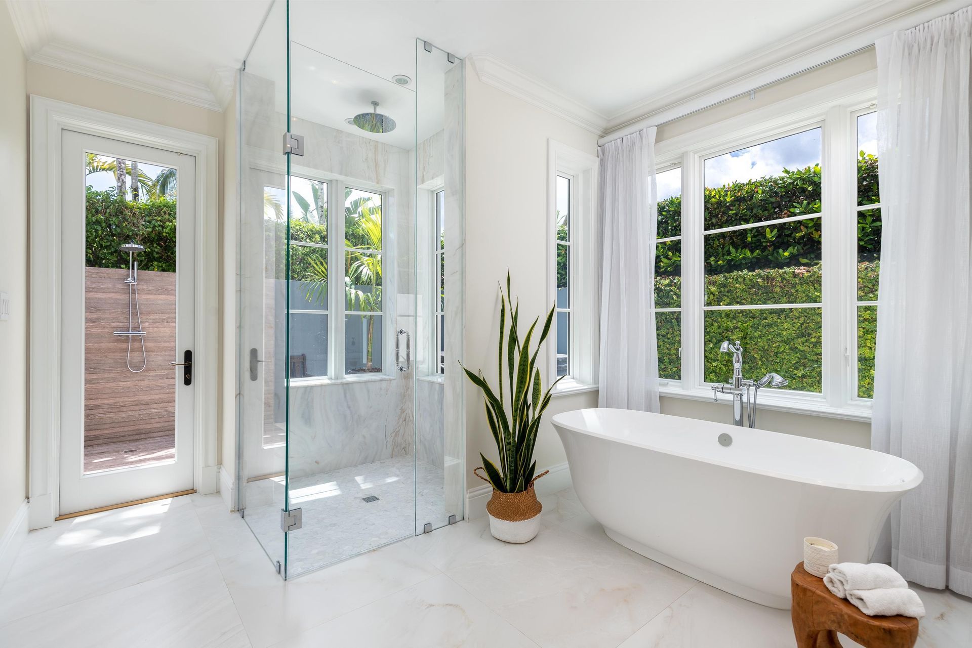 Bright white bathroom with a glass shower, soaking tub, and windows with greenery views.