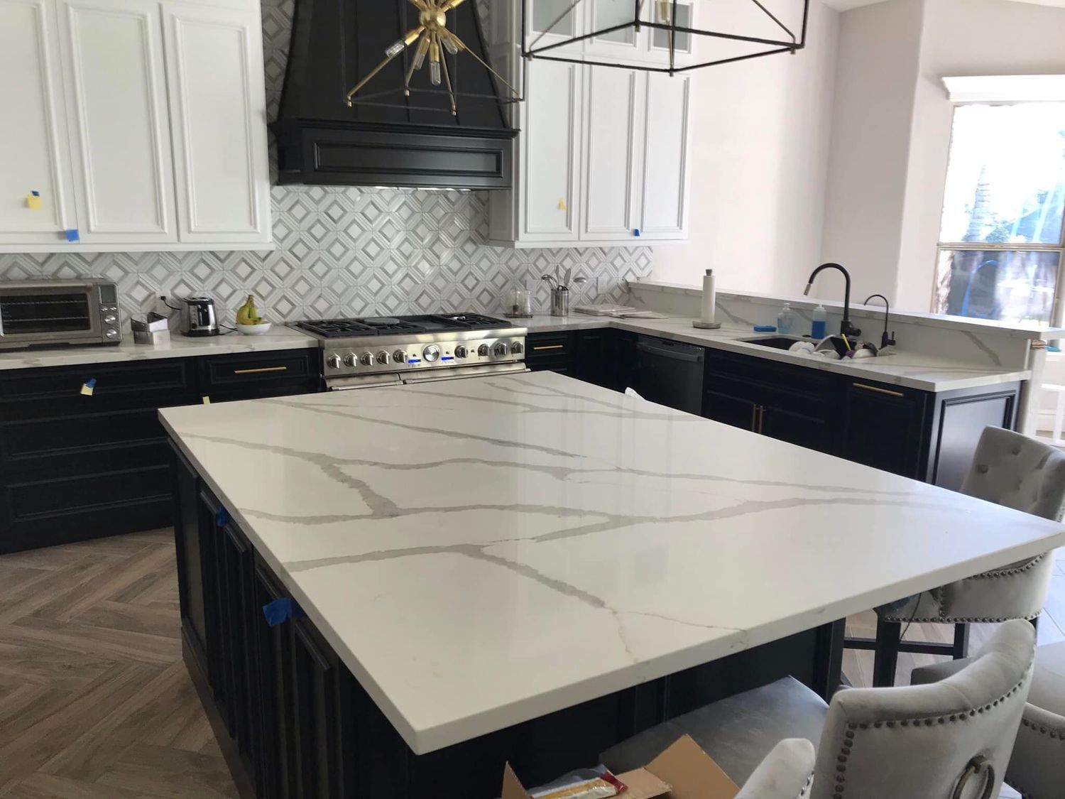 Kitchen with black and white cabinets, large island with white marble-look countertop, stove, and patterned backsplash.