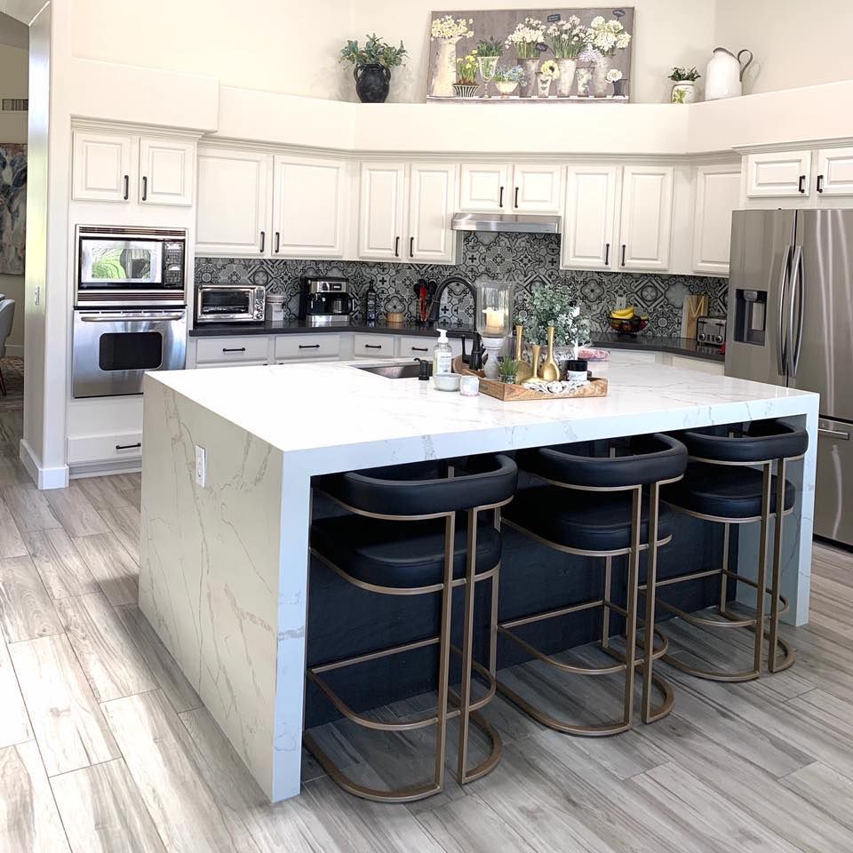 A modern kitchen with a large island and black and gold bar stools. White cabinets and a stainless steel fridge.