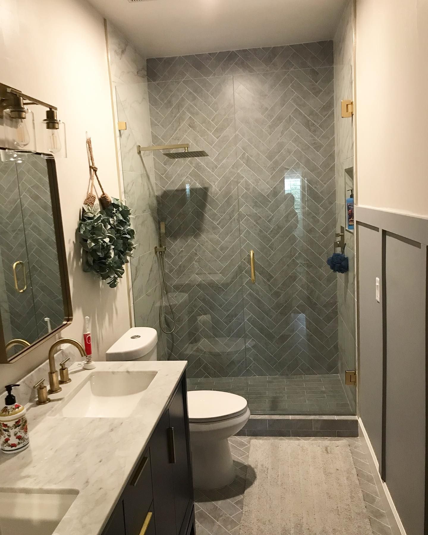 Bathroom with grey herringbone tile shower, marble vanity, and gold fixtures.