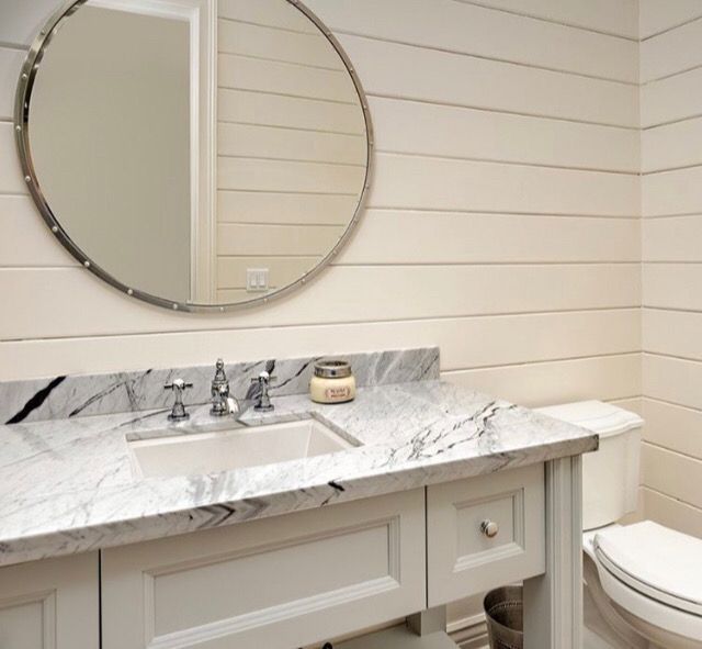 White bathroom with a round mirror over a marble countertop and shiplap walls.