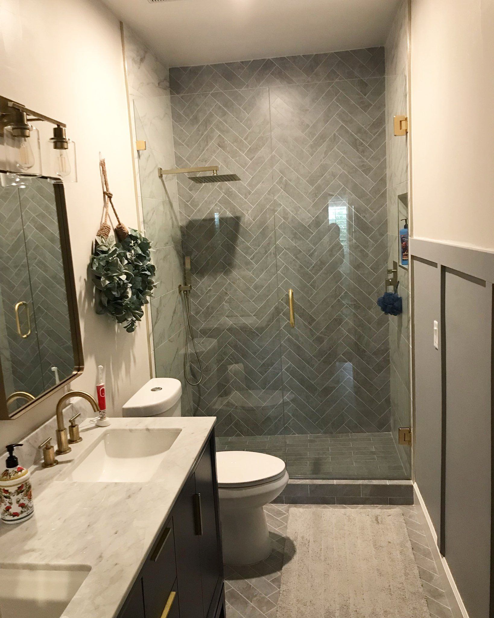 Bathroom with marble countertop, gray herringbone tile, gold fixtures, and glass shower.