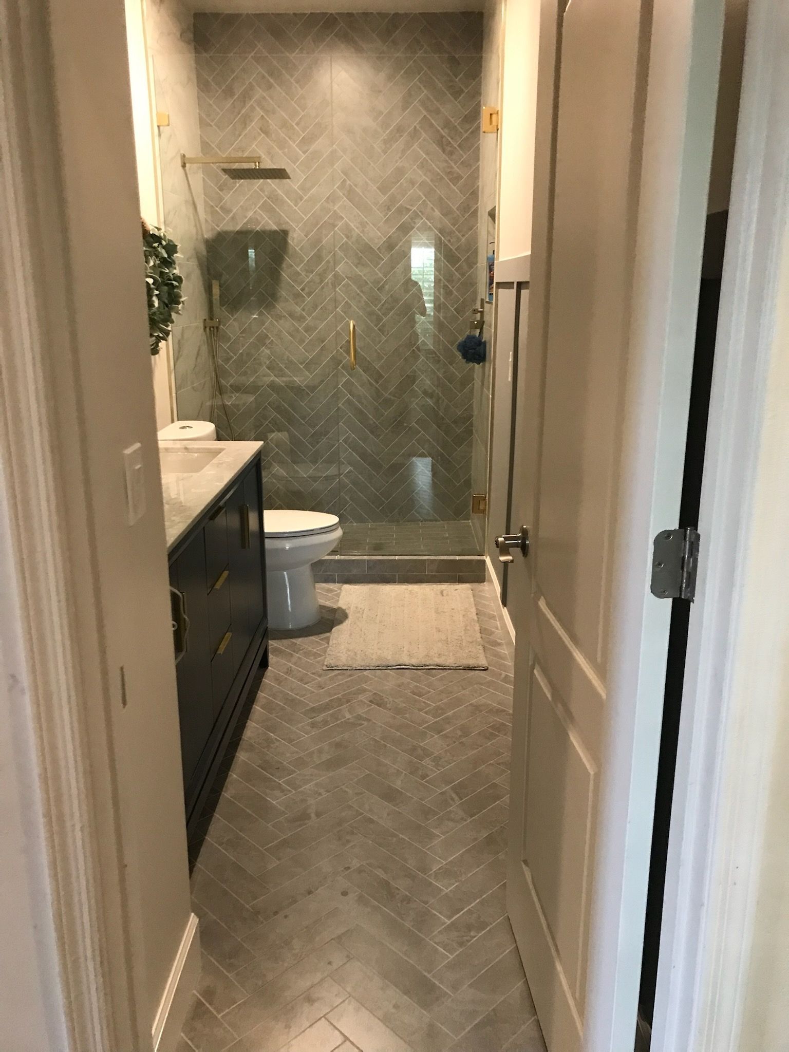 Narrow bathroom with herringbone tile floor, shower, toilet, and dark vanity.