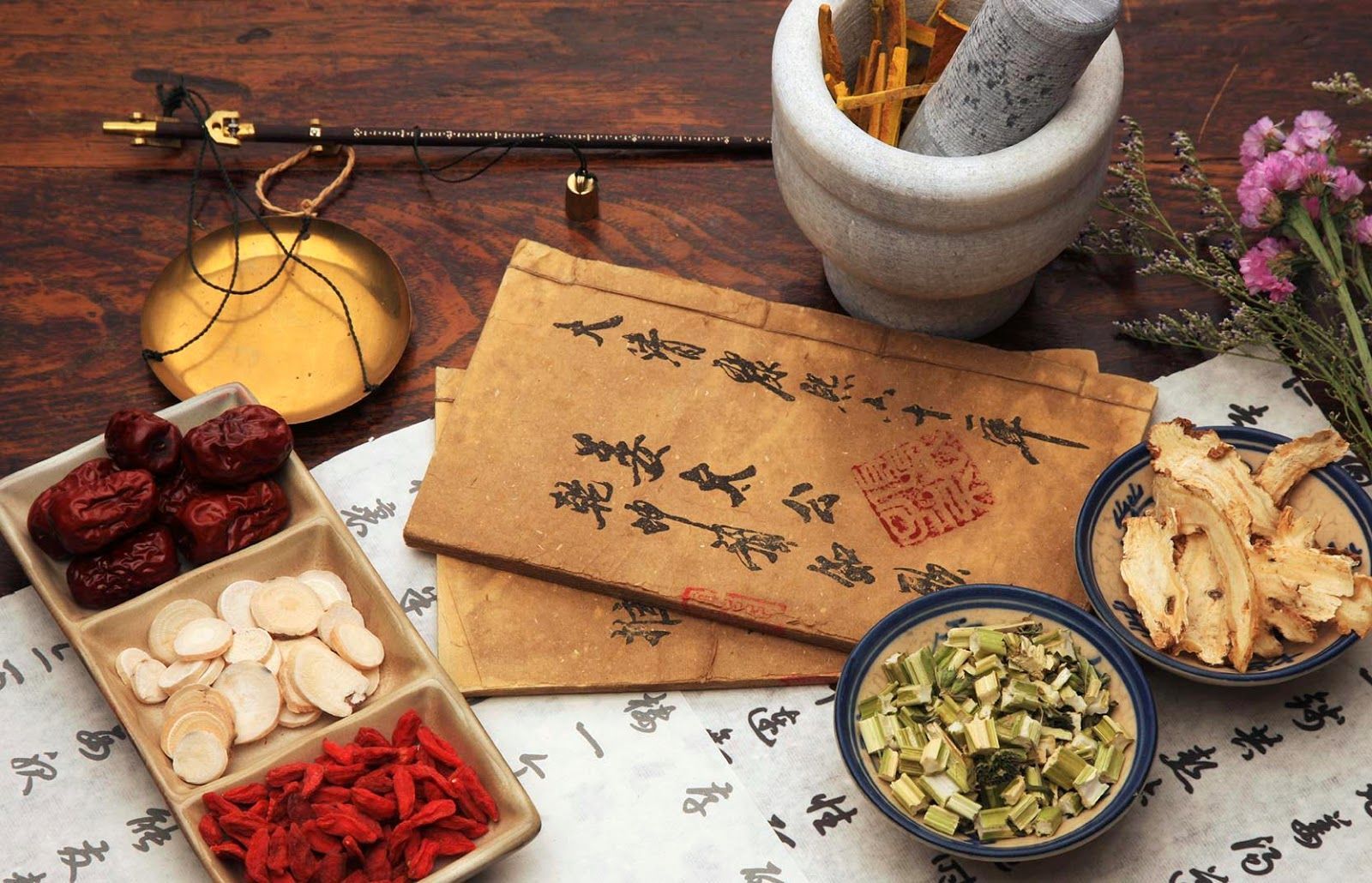 Woman with slicked-back hair and hoop earrings, wearing a beige and white top, with glittery skin, in front of an orange wall.
