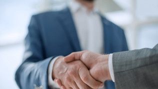 Two men shaking hands, one in a blue suit, the other in gray, likely sealing a deal indoors.