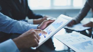 Person in a blue shirt points at charts on a tablet, surrounded by people, in a business setting.