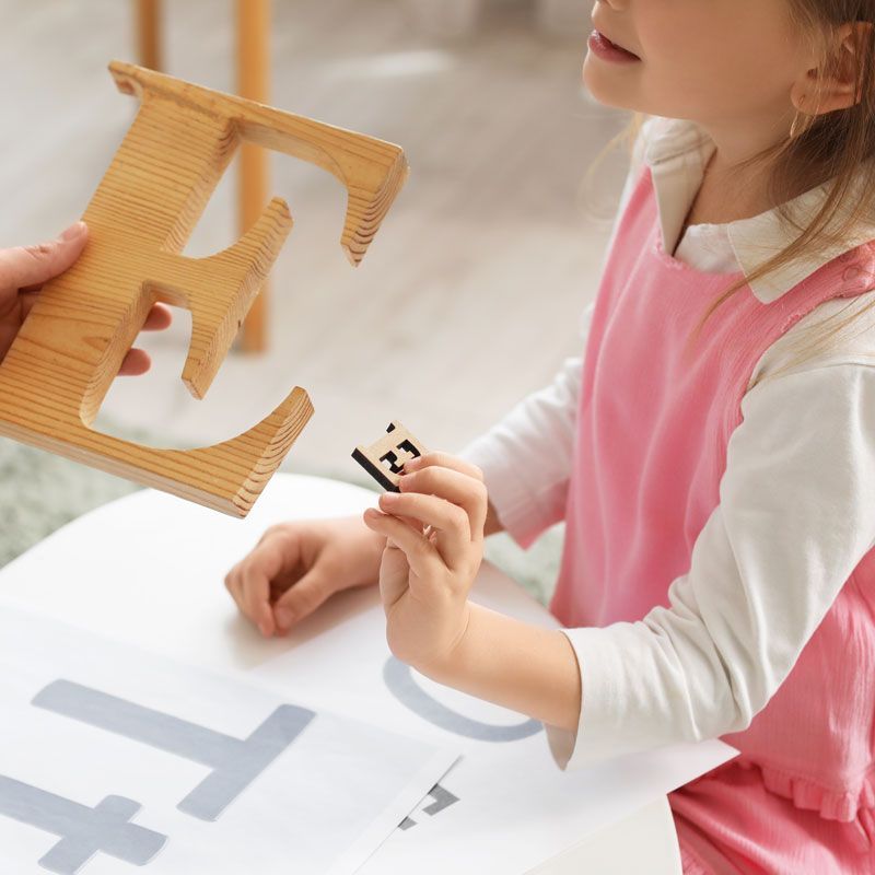 A little girl in a pink dress is holding a wooden letter e