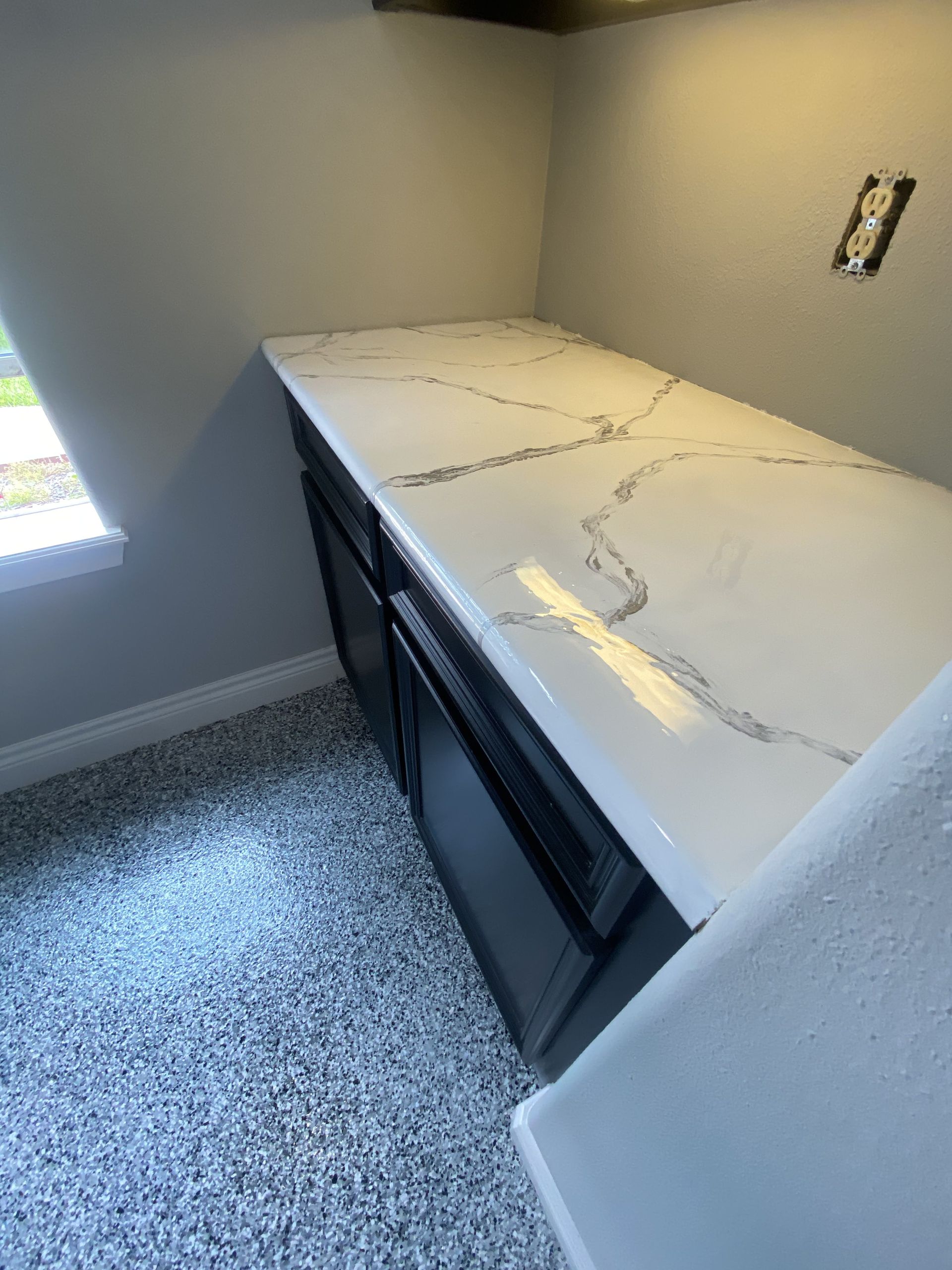 White countertop with gray veining on dark cabinets, gray walls, and speckled flooring.