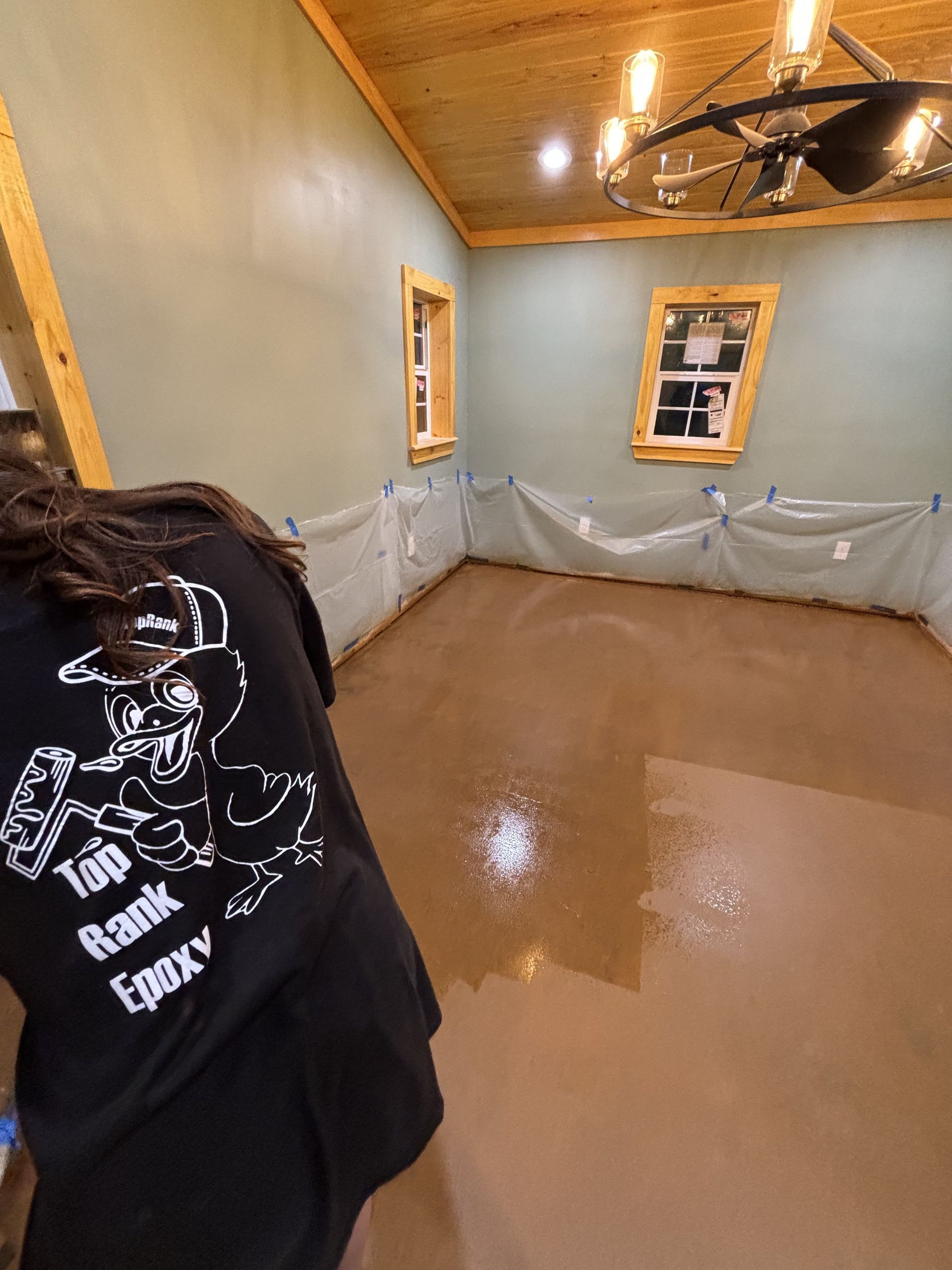 Person applying epoxy to a brown floor in a room. Walls are green with plastic sheeting taped along the bottom.