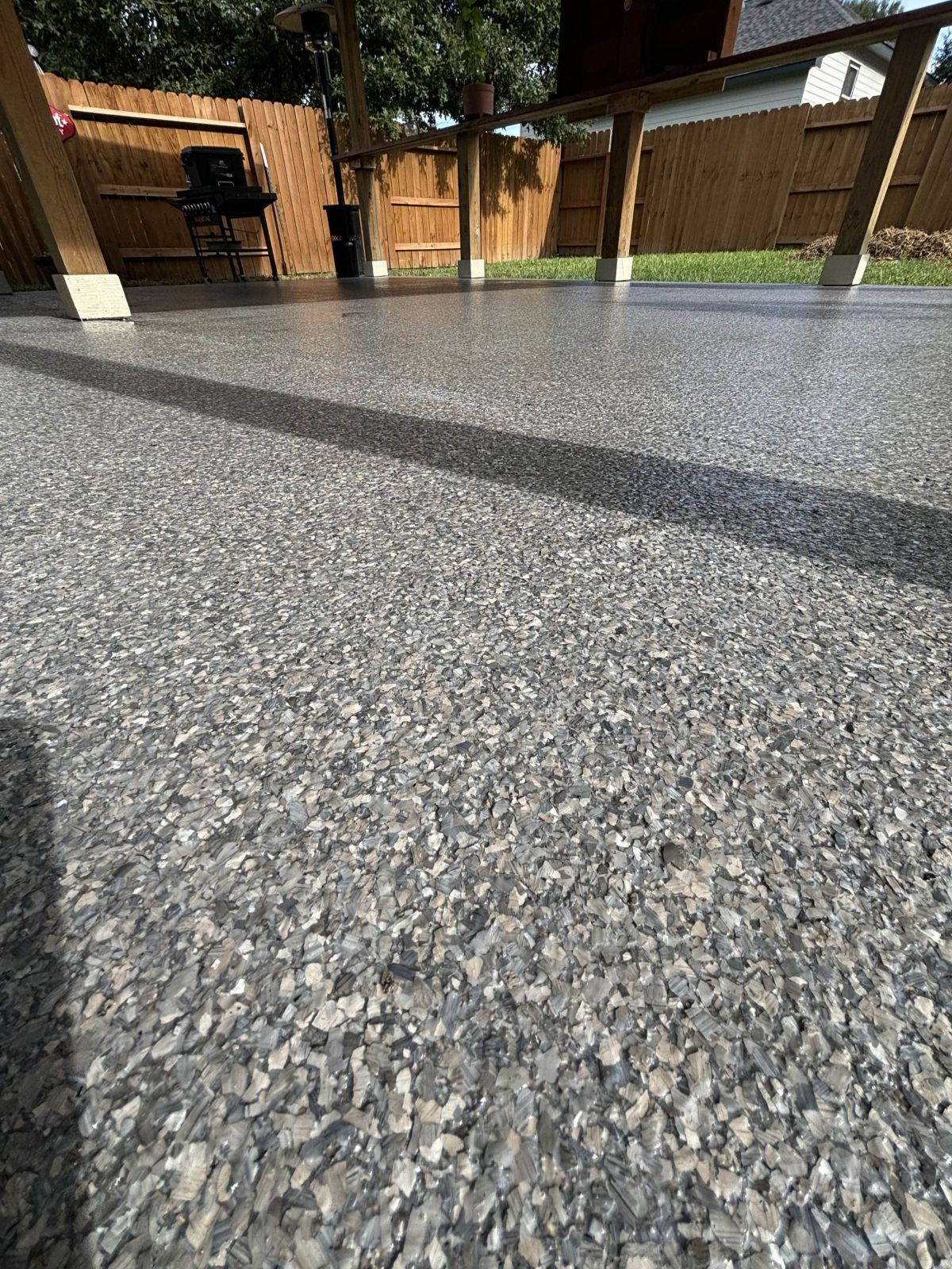 A gray speckled outdoor concrete patio with a wooden fence and structure in the background.