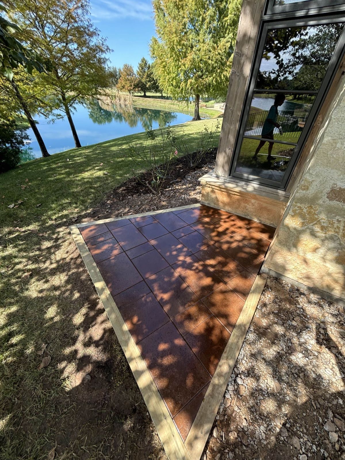 Triangular patio with dark tiles next to a building and a lake with trees in the background.
