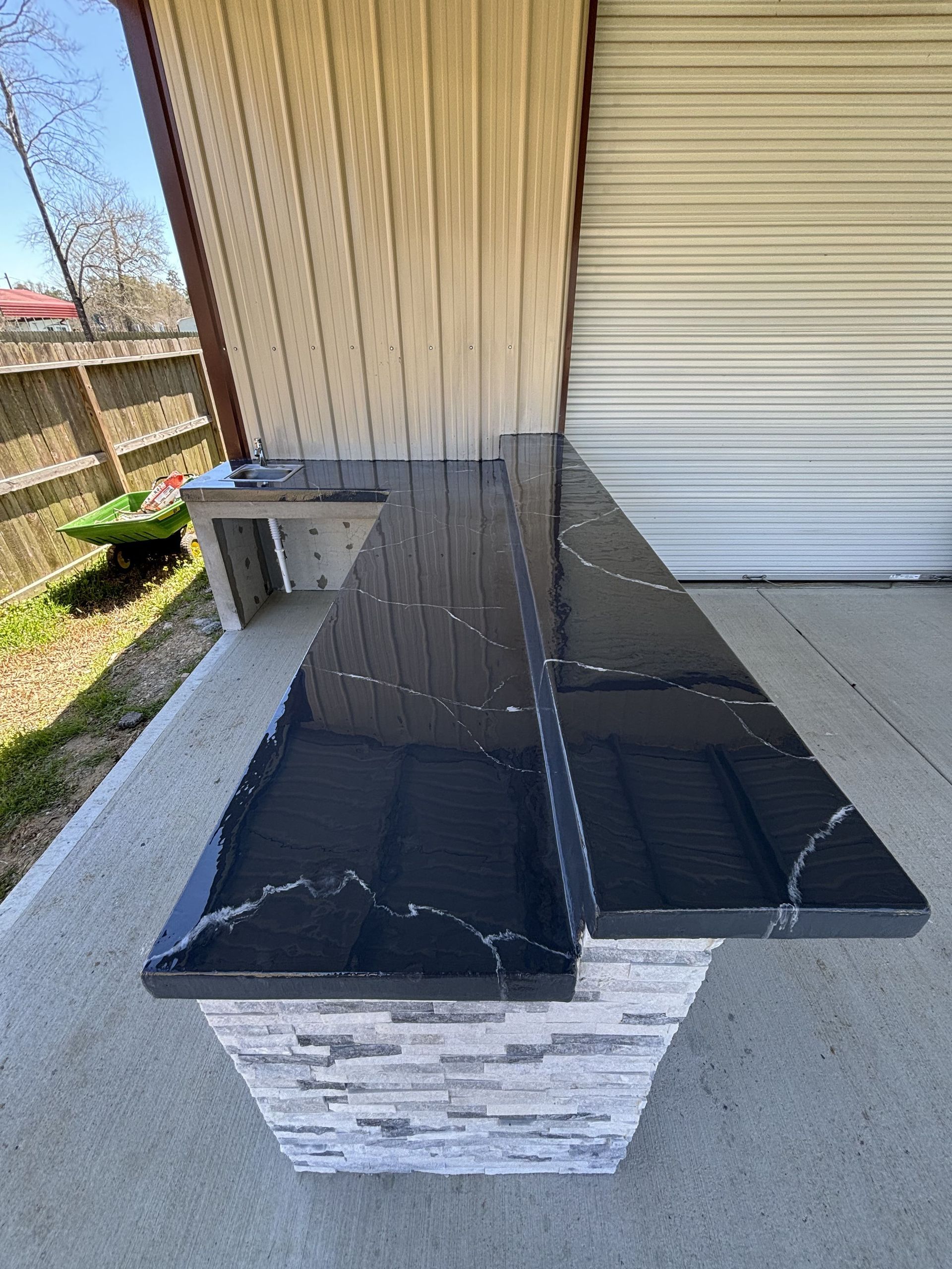 Outdoor bar with black countertop and stone base, next to a garage door.