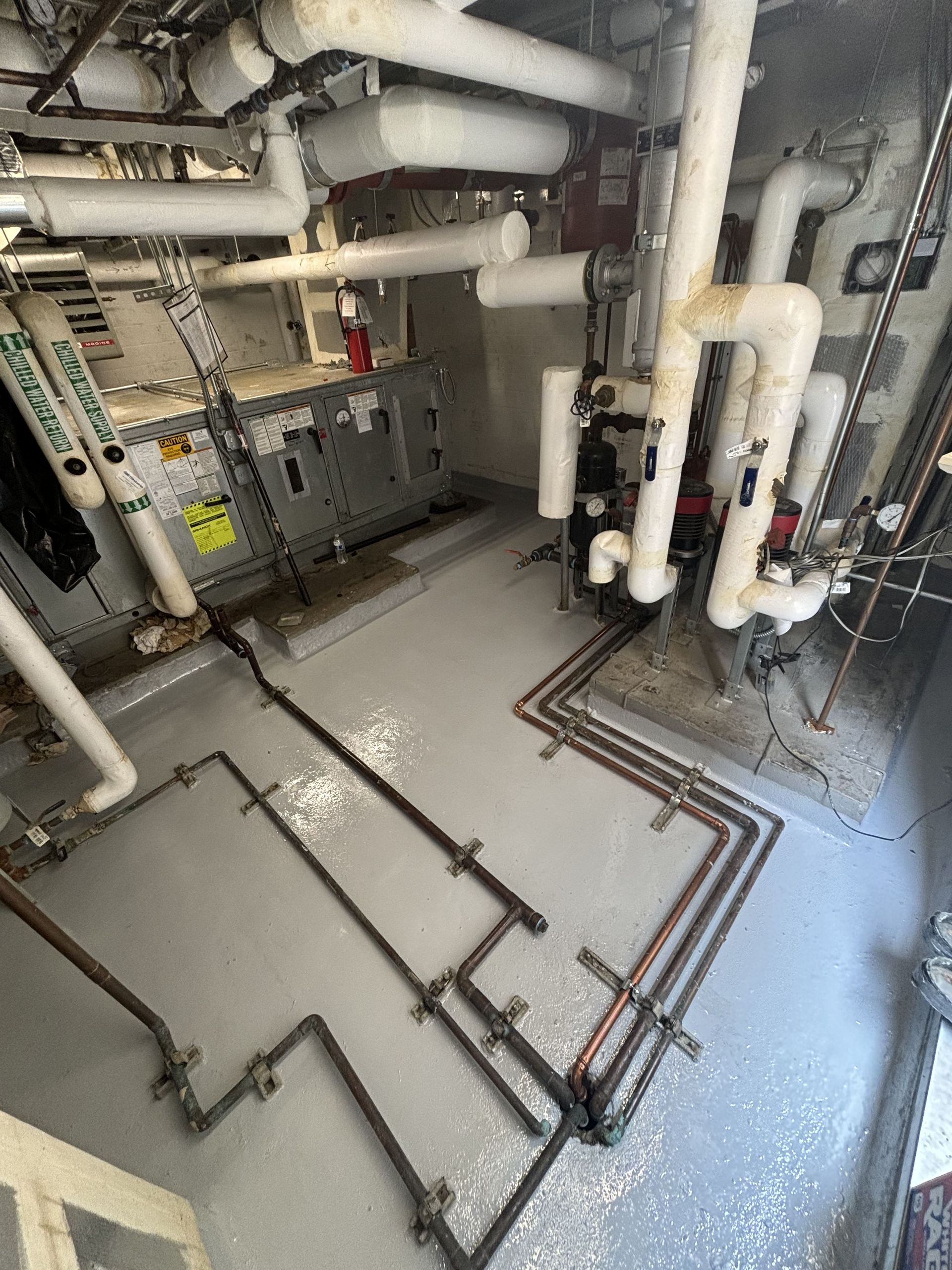 A mechanical room with pipes, equipment, and a gray floor. White insulation covers some pipes.