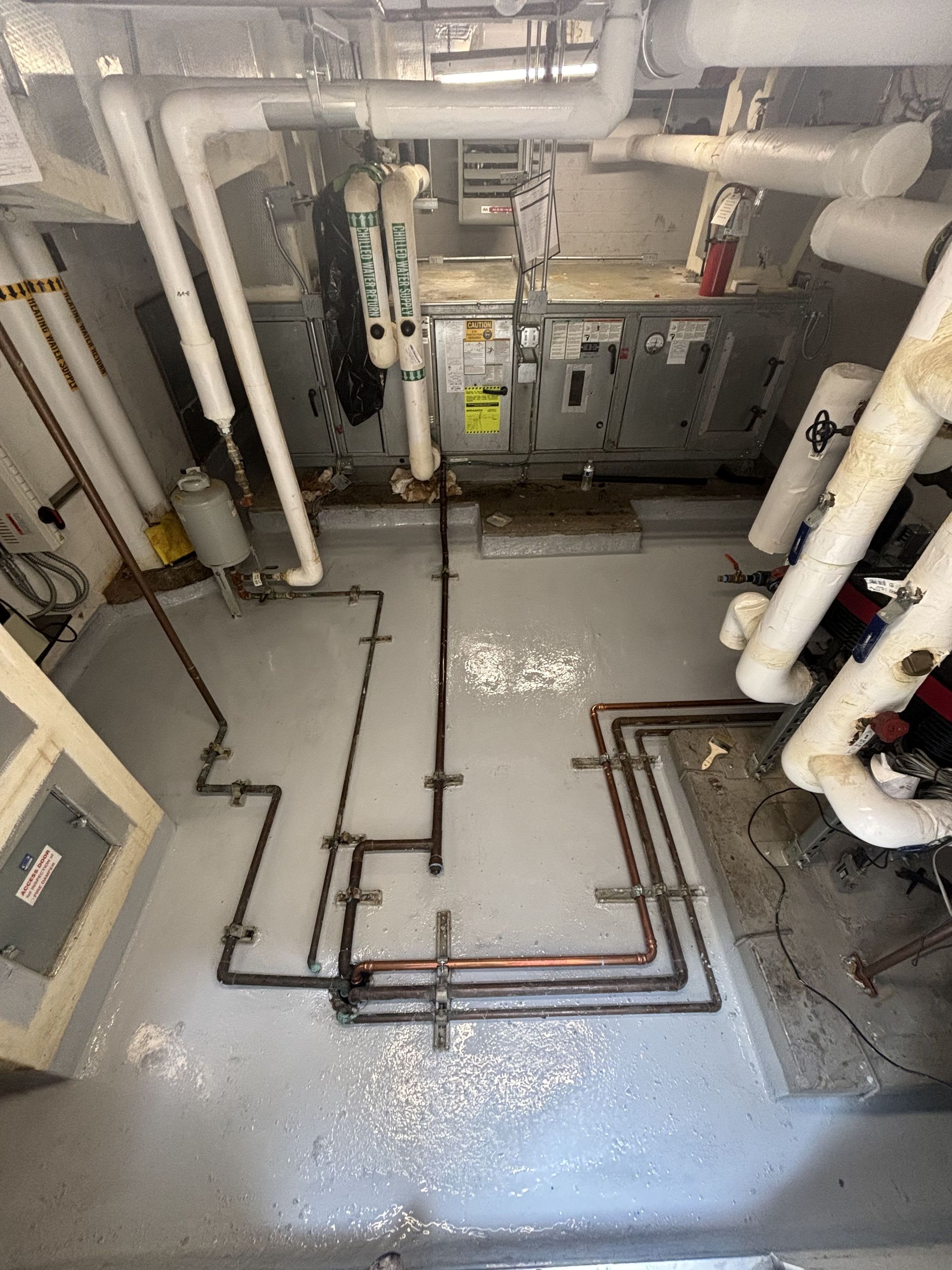 Mechanical room with pipes, HVAC equipment, and wet floor. White and copper pipes, silver machinery.