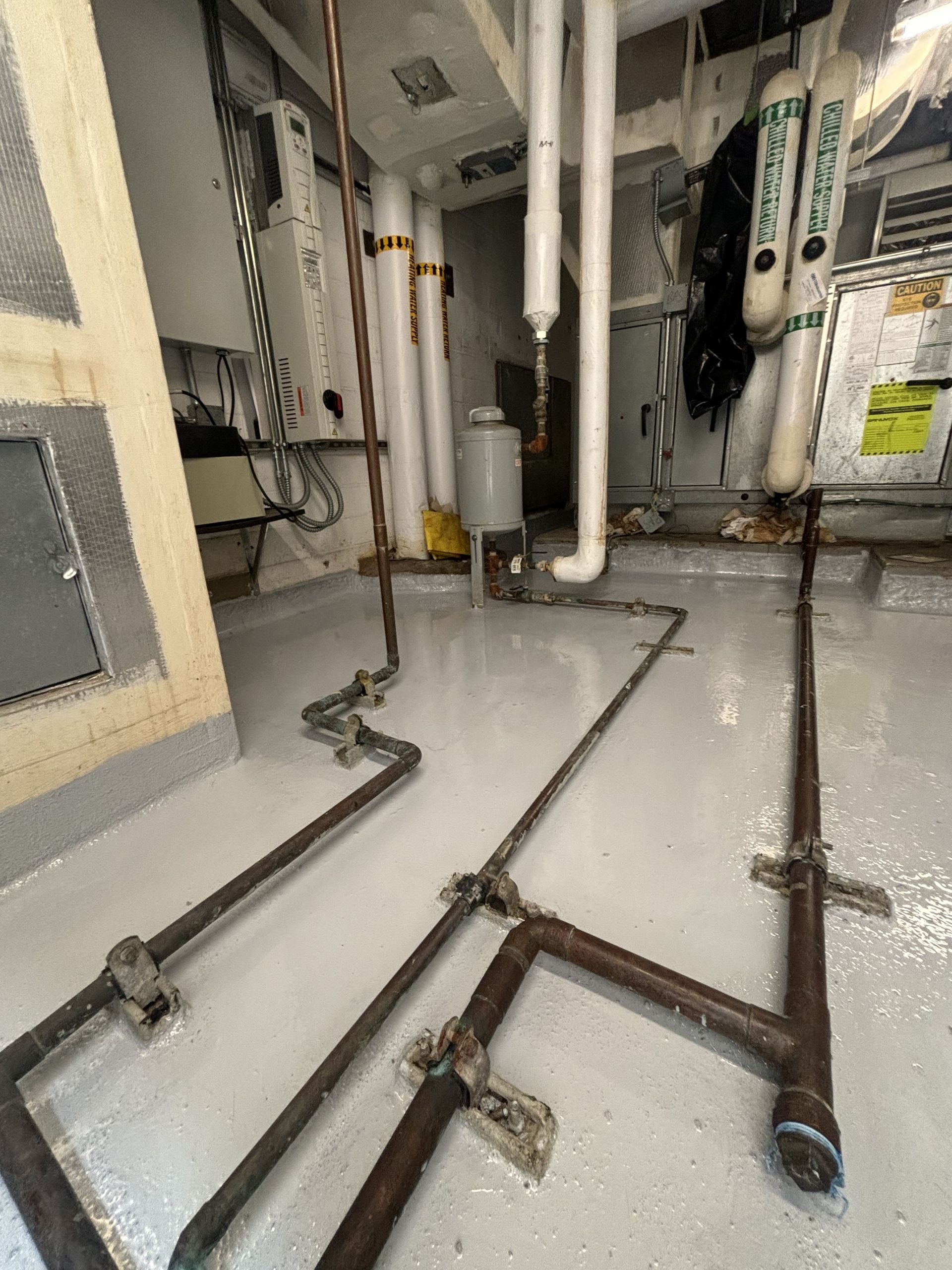 Pipes and equipment in a utility room with white flooring. Copper and white pipes run along the floor and walls.