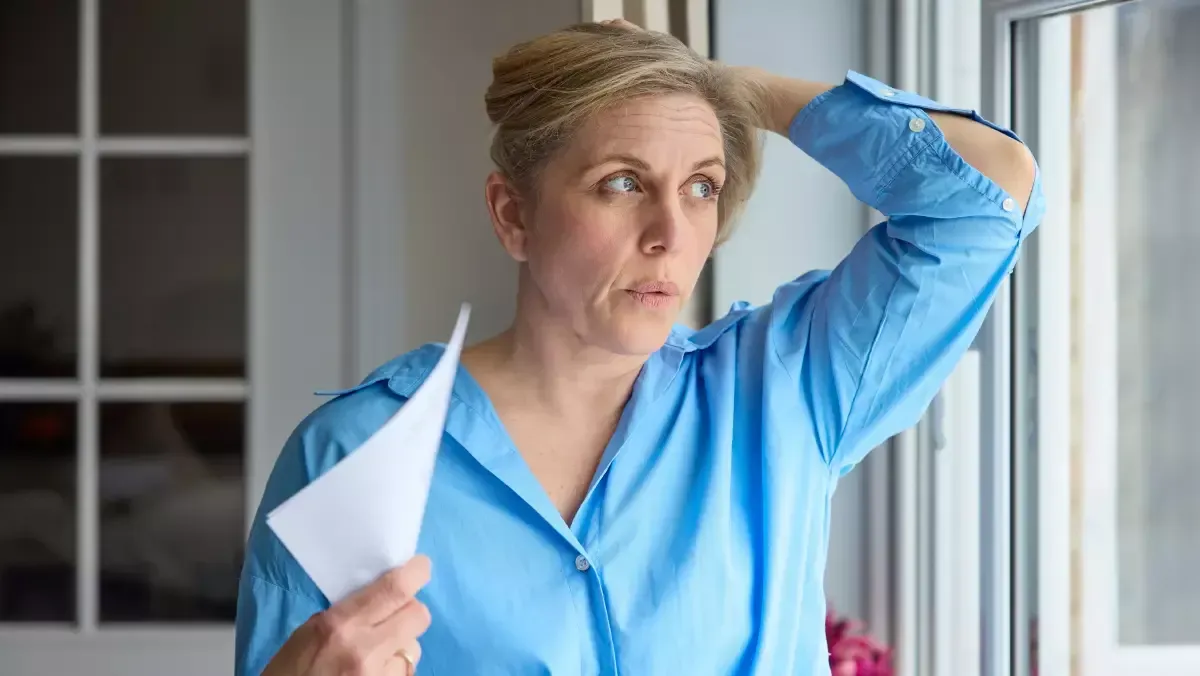 A woman in a blue shirt is standing in front of a window holding a piece of paper.