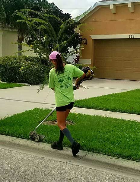 A woman is walking down the sidewalk while using a lawn mower.