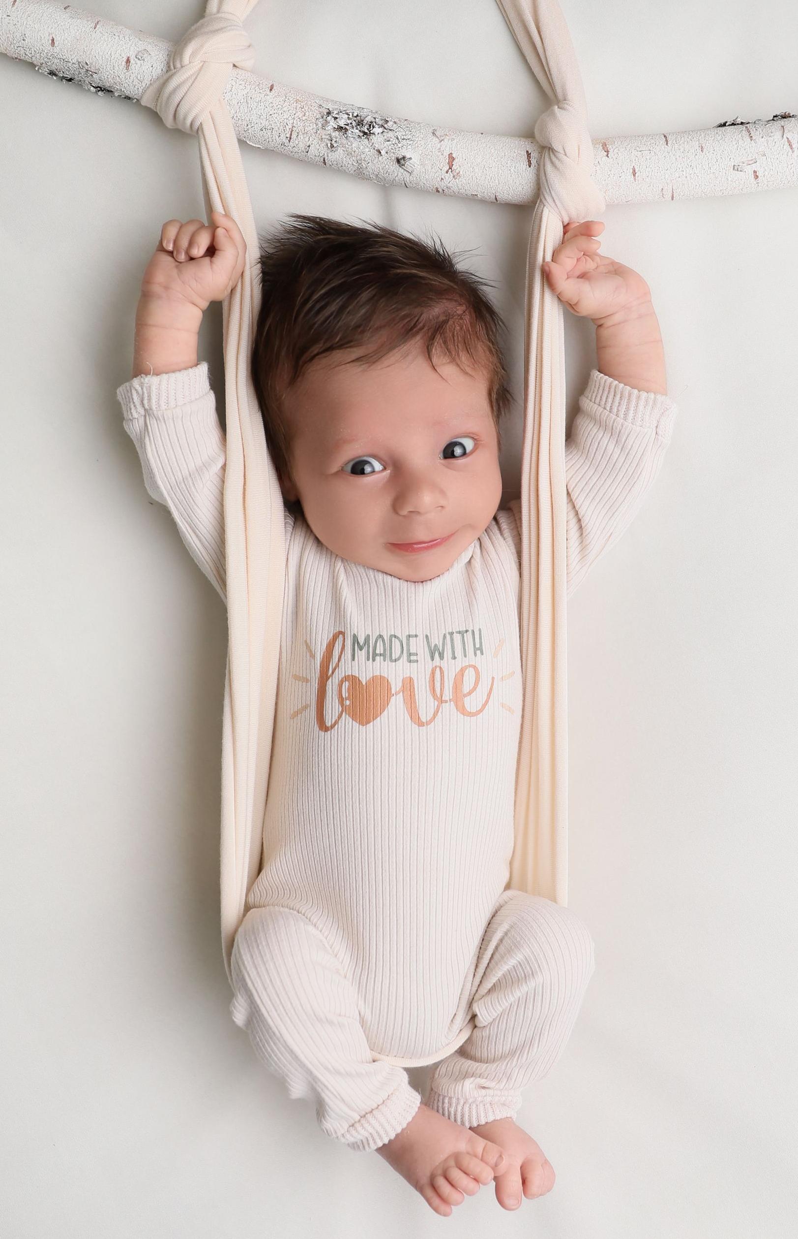 An awake newborn lies comfortably on a soft, white posing surface, gently supported with neutral fabric straps for safety. The baby’s wide, curious eyes are open, capturing a calm and alert expression. Dressed in a simple cream outfit that reads “Made with Love,” the image highlights natural connection, security, and the beauty of an awake newborn moment during a professional studio session.