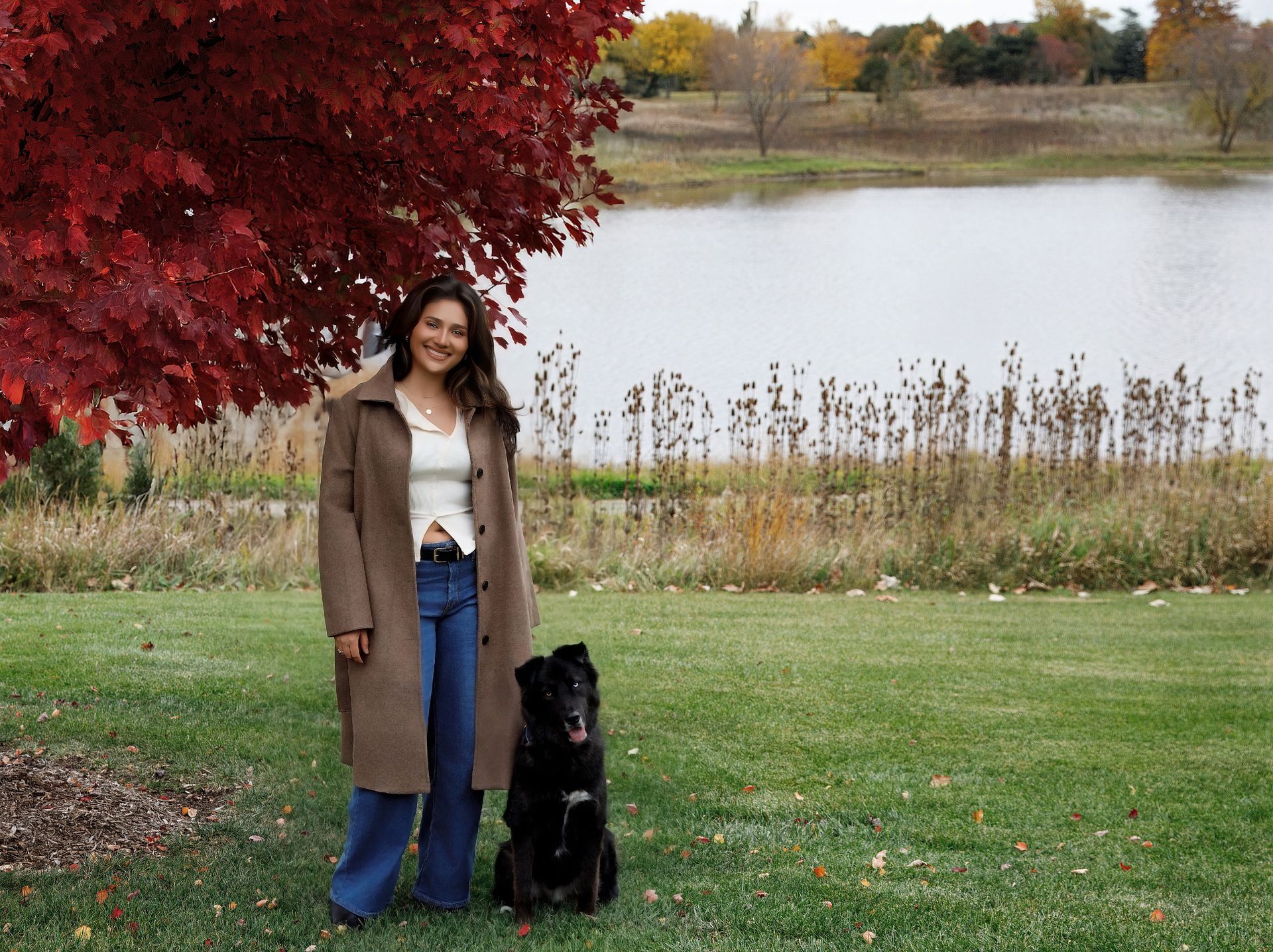 Woman in brown coat and jeans with black dog under a tree with red leaves, by a lake.