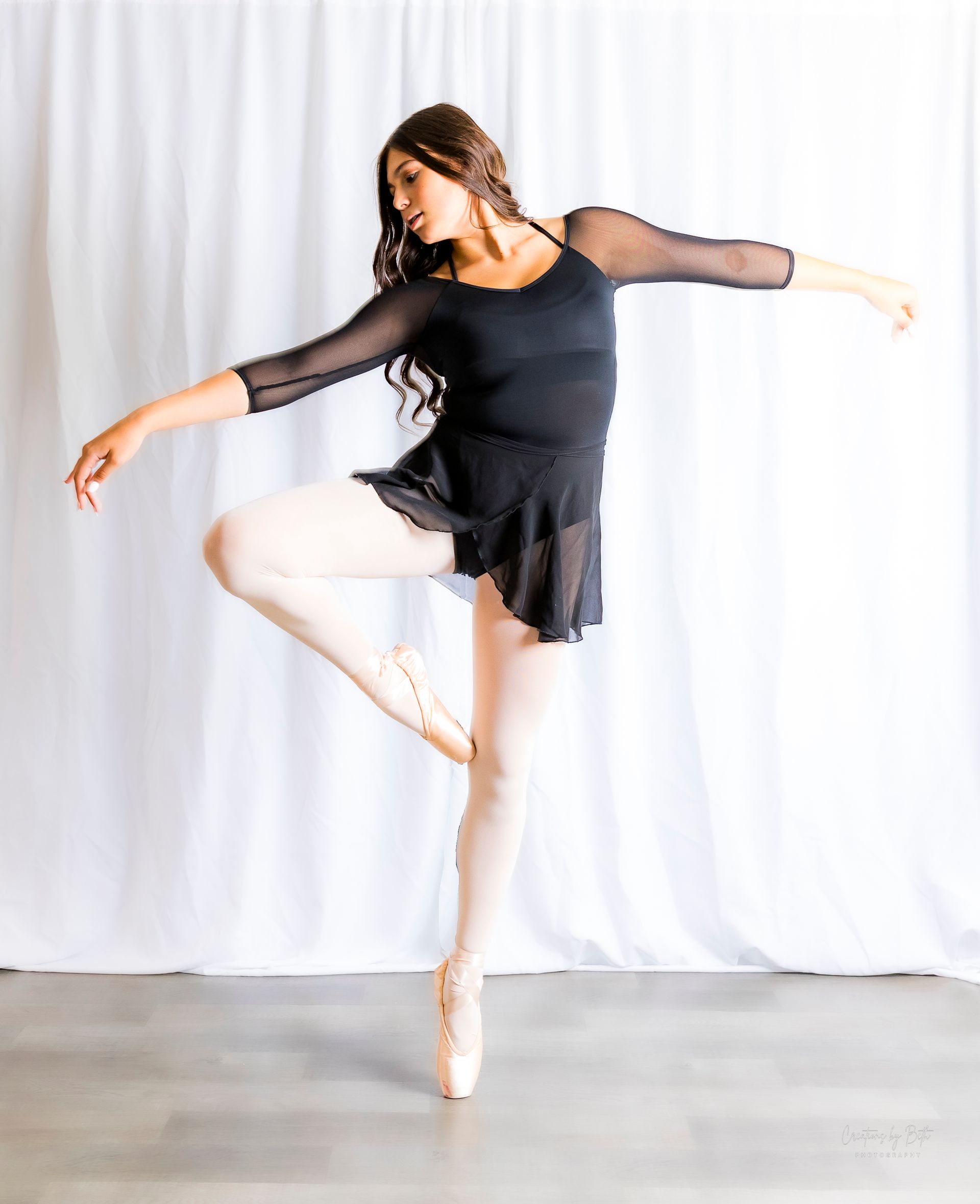 A woman in a black dress and pointe shoes is dancing in front of a white curtain.