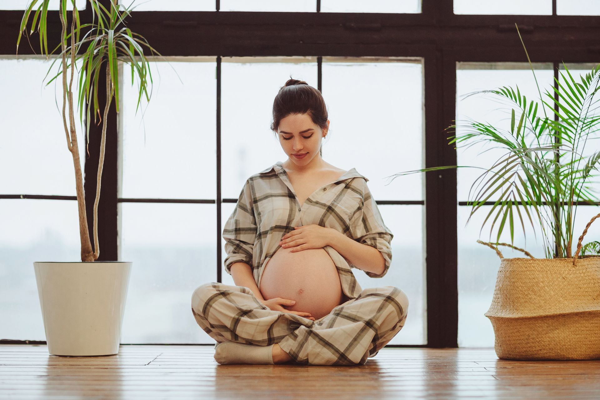 A pregnant woman is sitting on the floor in front of a window holding her belly.