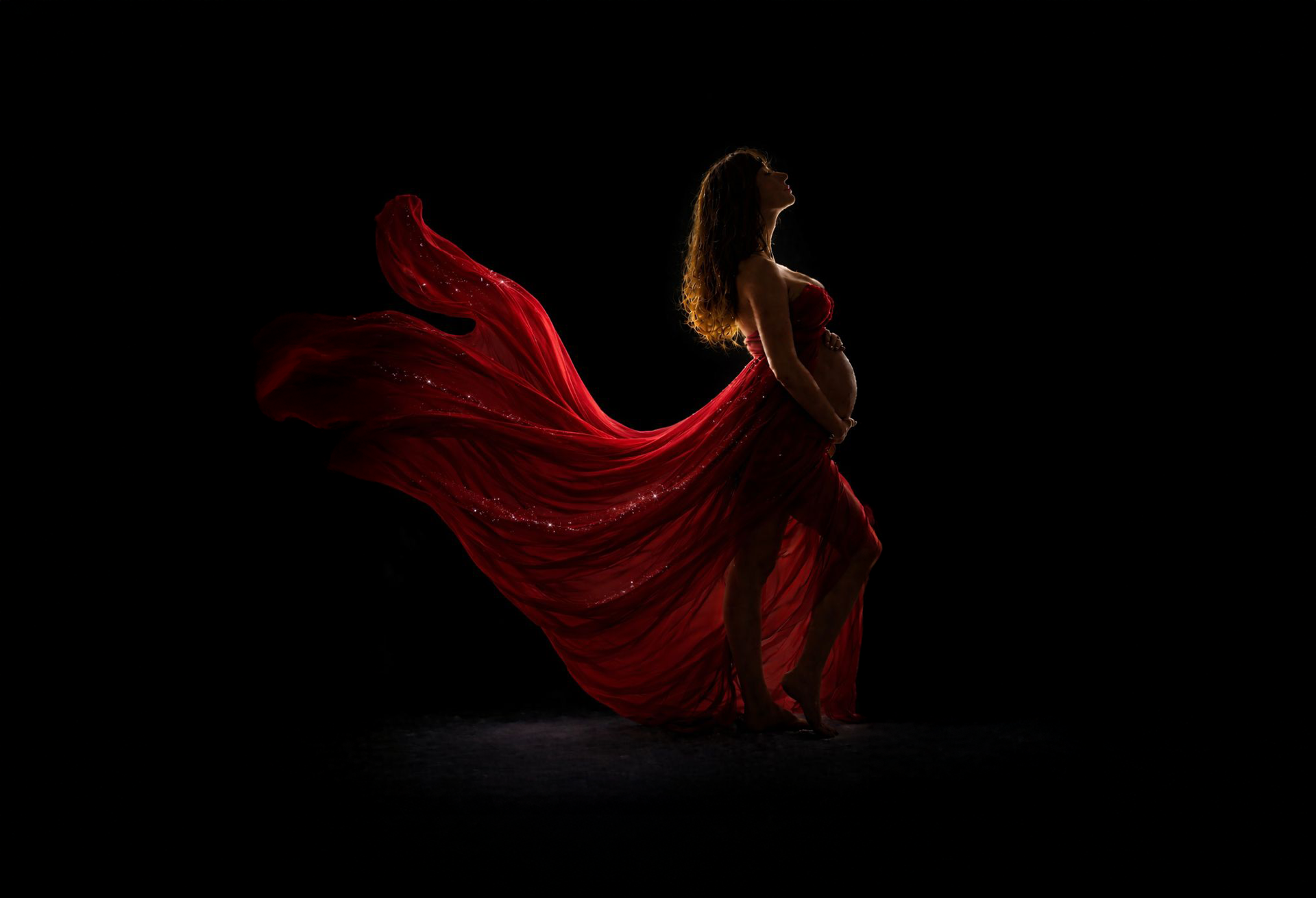 Pregnant person in silhouette, wearing red fabric, looking upward.