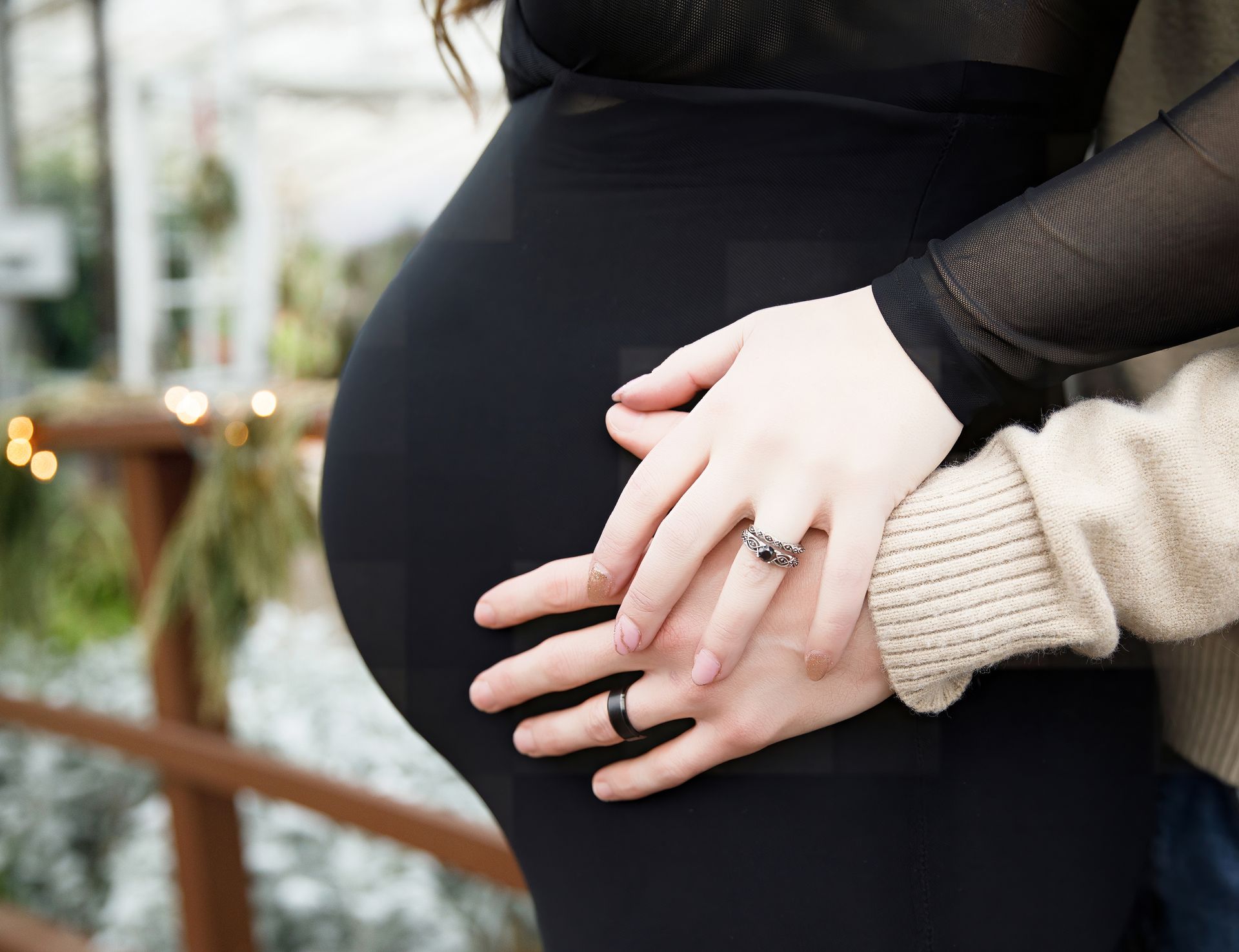 Pregnant person in a black dress, hands gently resting on belly, ring visible. Outdoor setting with greenery.