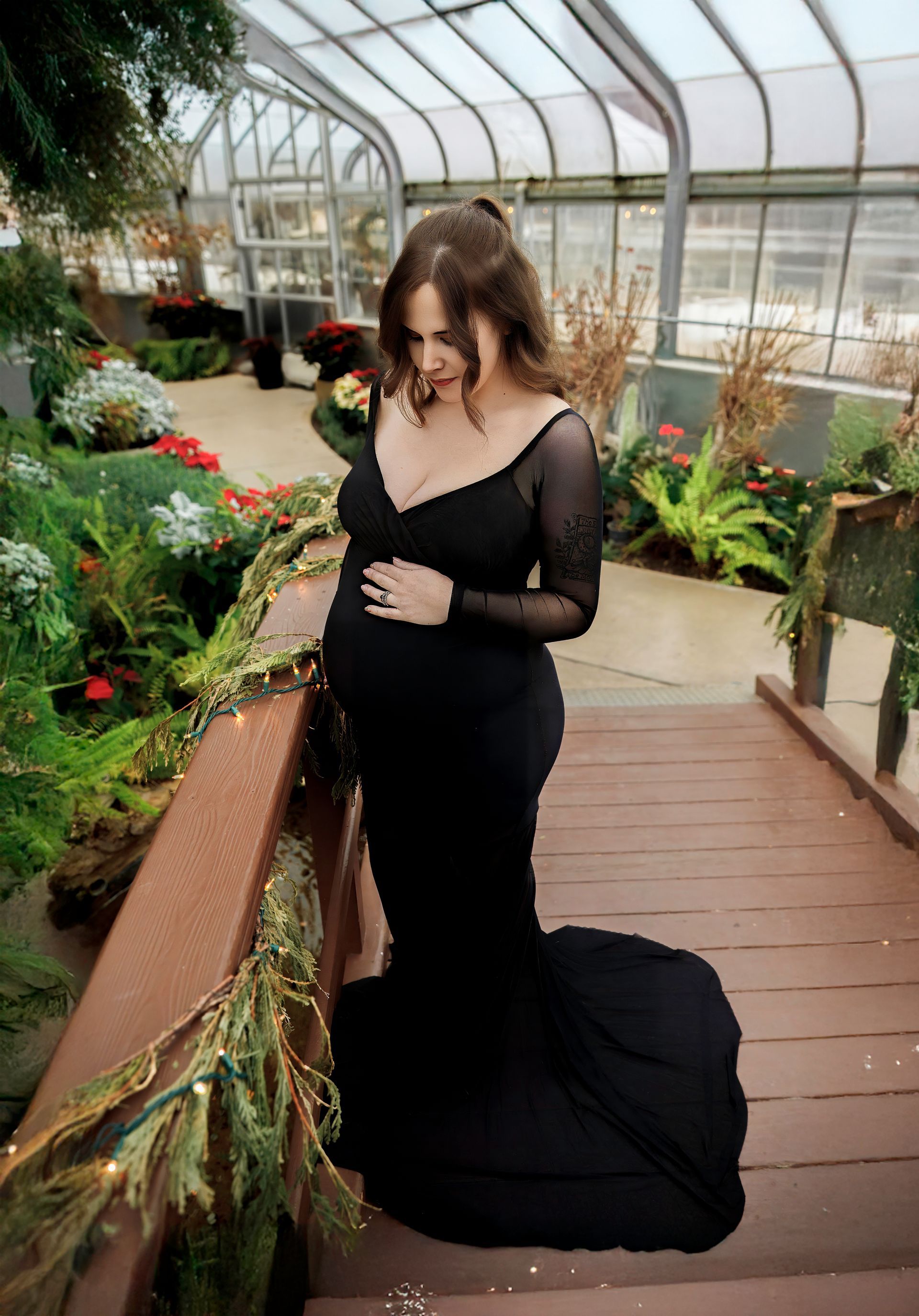 Pregnant person in a black gown poses on a wooden walkway in a greenhouse, touching their belly.