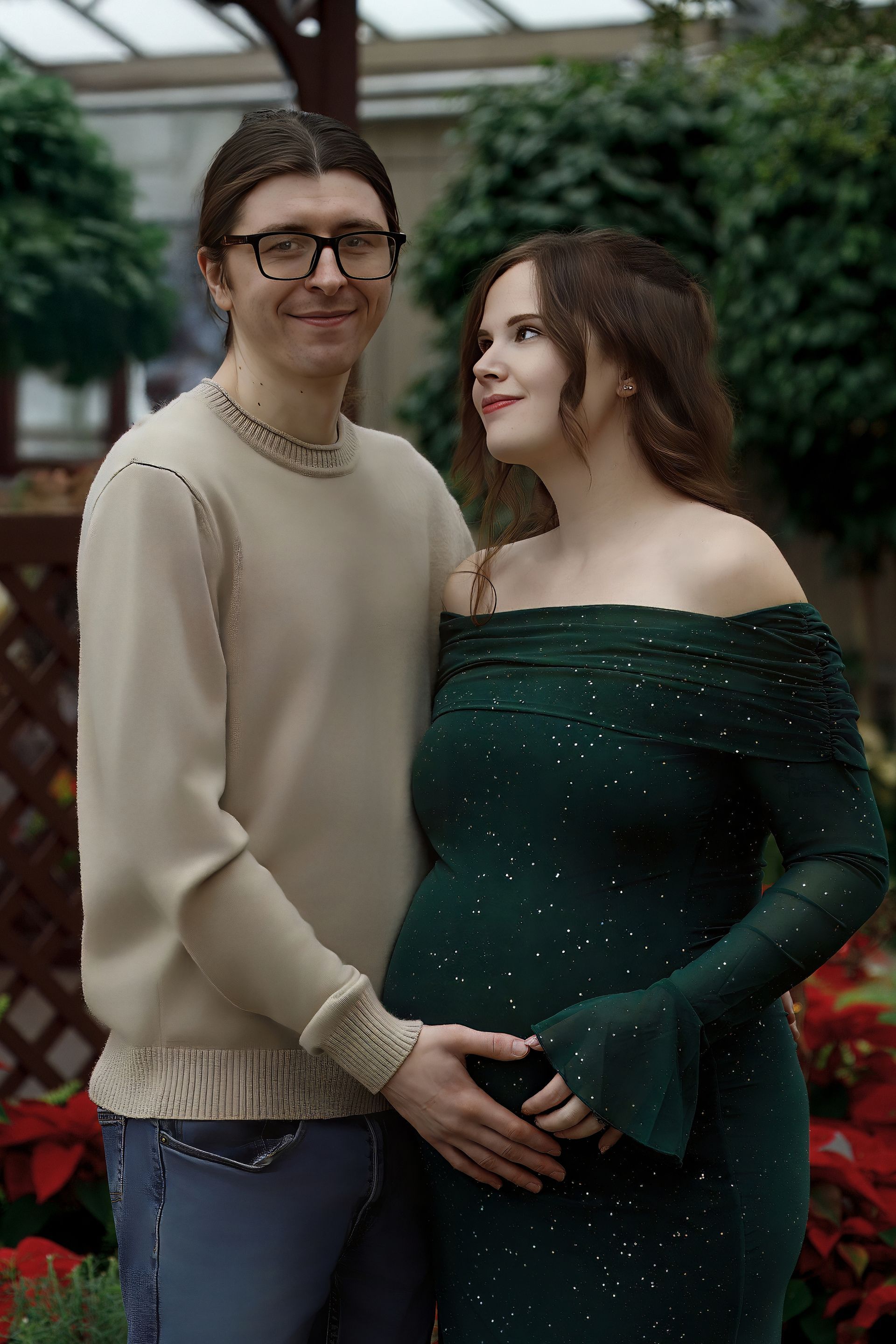 Couple holding pregnant woman's belly. Man wears glasses and sweater. Woman in green dress. Green foliage in background.