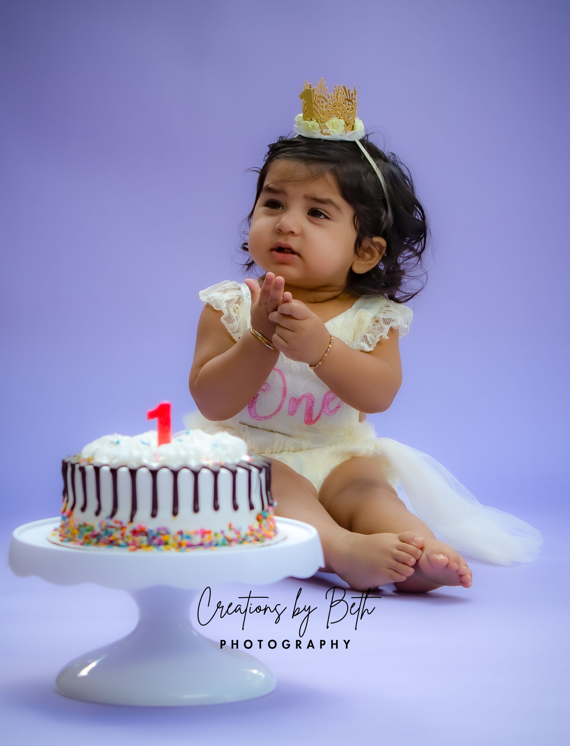 A baby girl wearing a crown and a white lace outfit sits near a cake with a