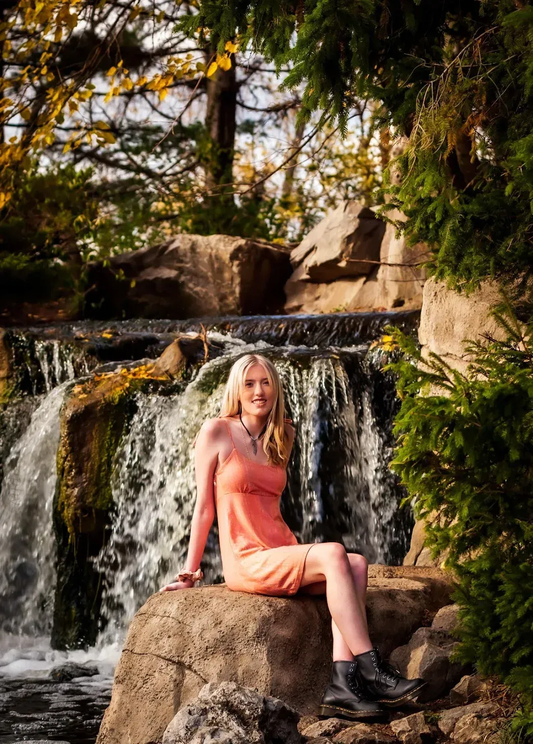 Woman in orange dress sits on a rock near a waterfall. She's smiling, with blonde hair and black boots in a natural setting.