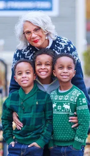 A woman is standing next to three young boys wearing green sweaters.