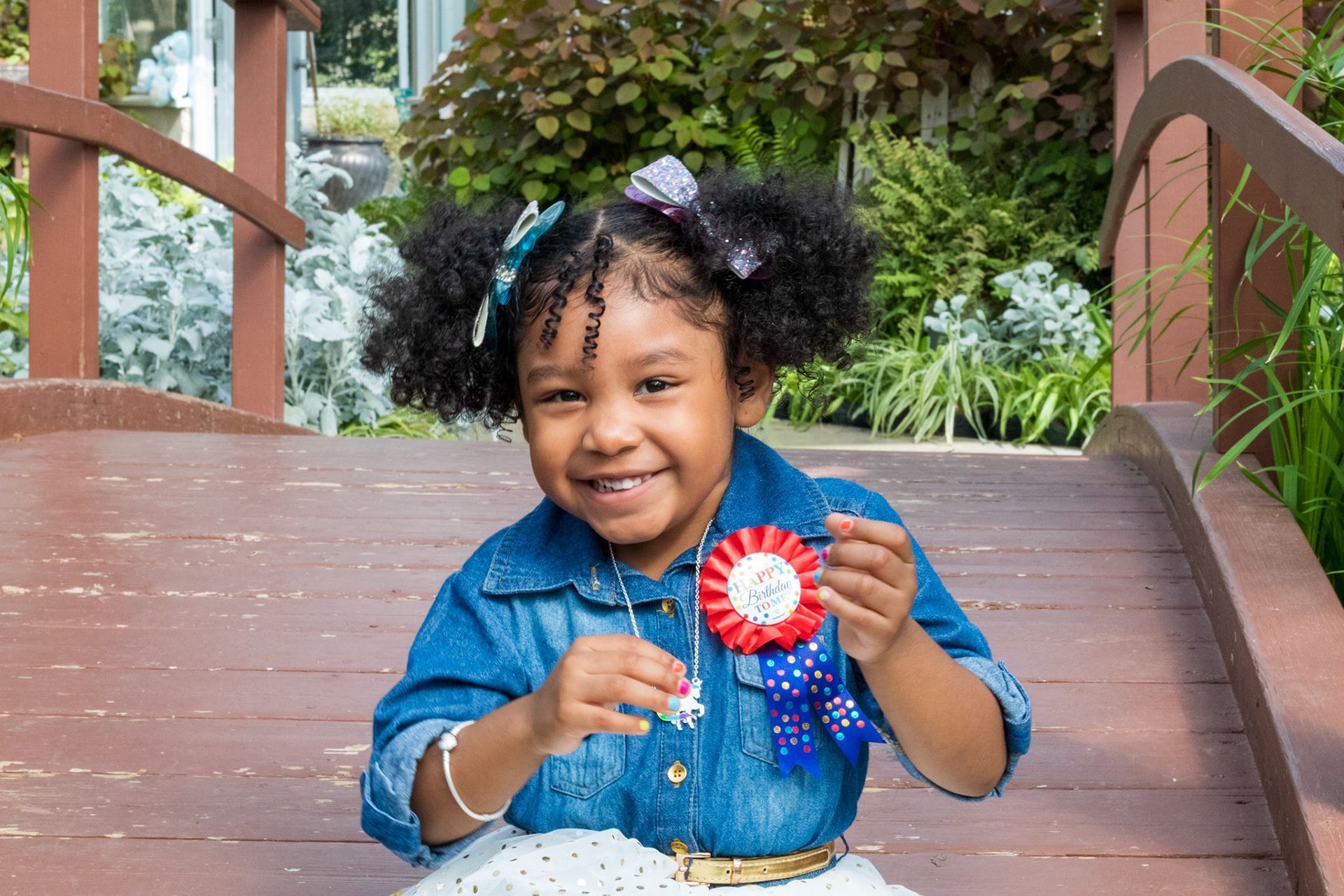 A little girl is sitting on a wooden bridge holding a red , white and blue ribbon.