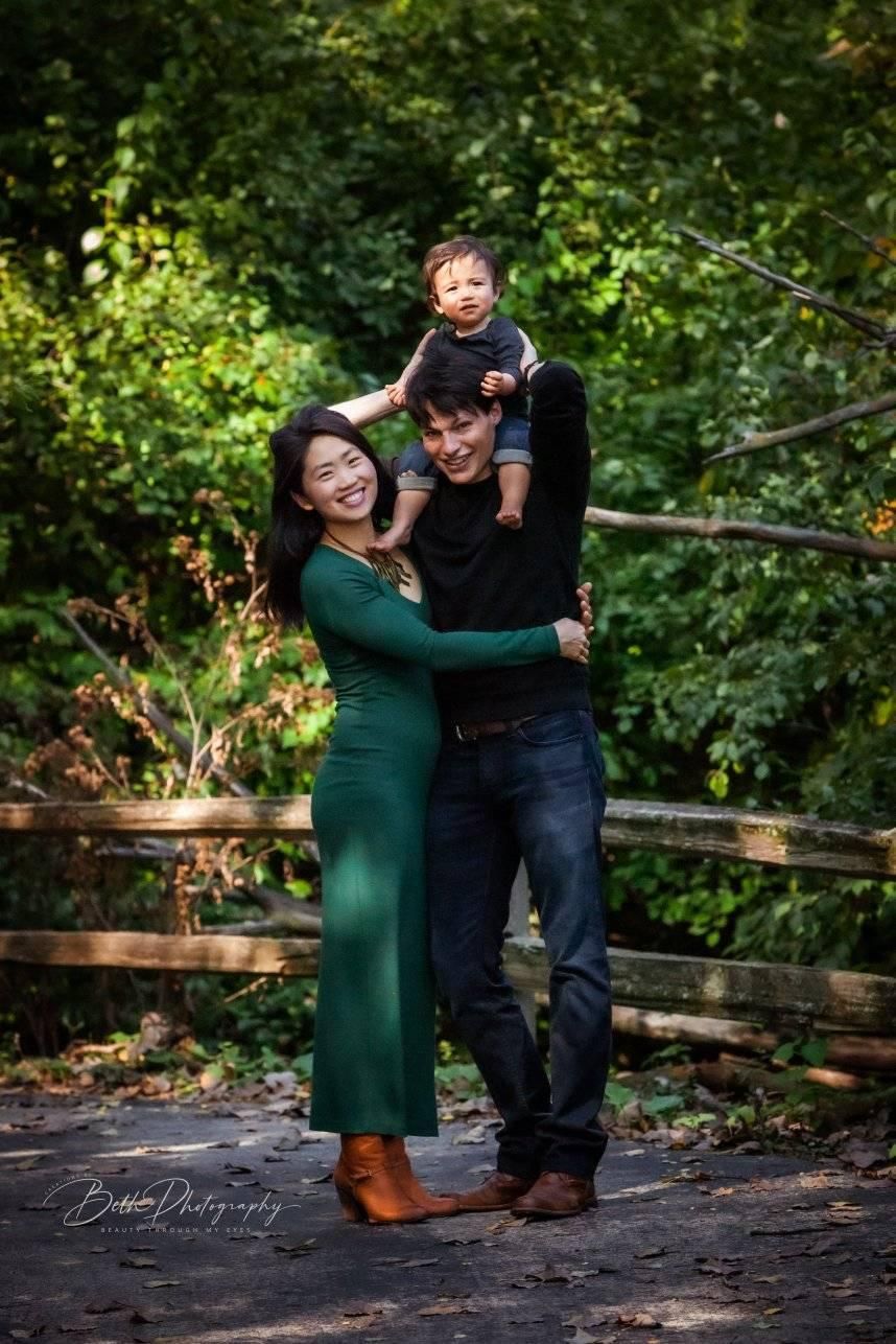 Family portrait. Mom in green dress and dad holding child on shoulders in a park with wooden fence and lush greenery.