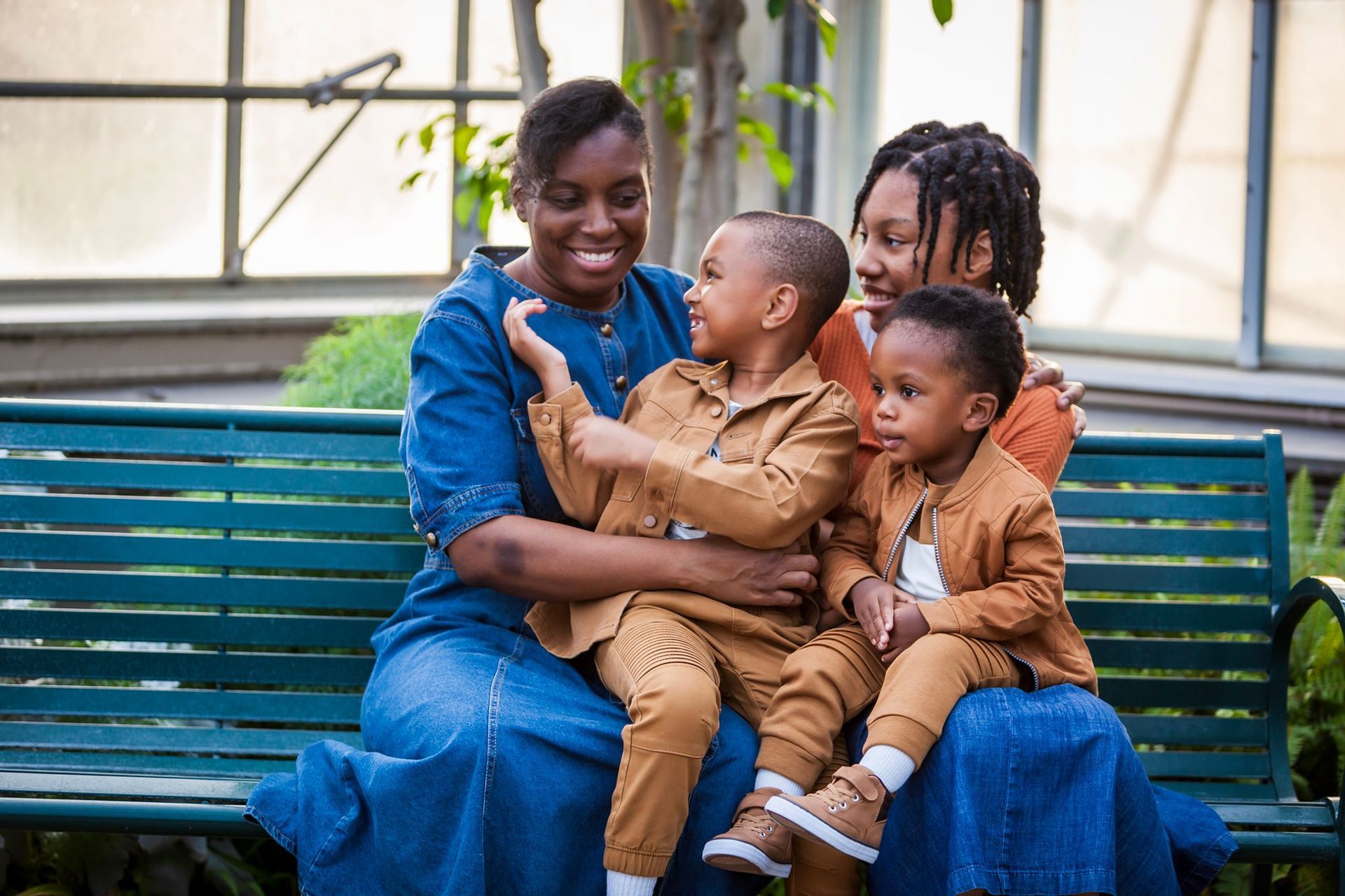 A smiling Black woman and two children sit on a bench, with a woman in dreadlocks next to them, in a sunny garden.