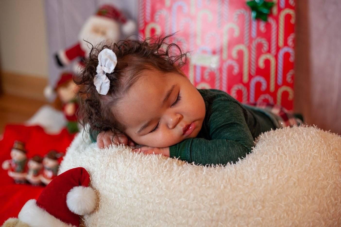 A sleeping baby rests on a soft, white surface in front of a Christmas scene with gifts and decorations.