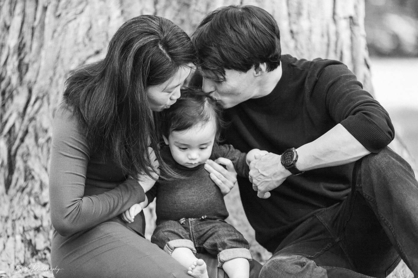 Family of three embracing by a tree. Parents, in dark clothes, kiss and cuddle their baby. Black and white.