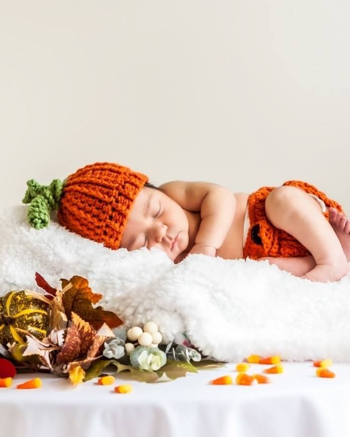 Newborn baby dressed in orange crochet pumpkin outfit, sleeping on a white fluffy blanket, with fall foliage and candy corn.