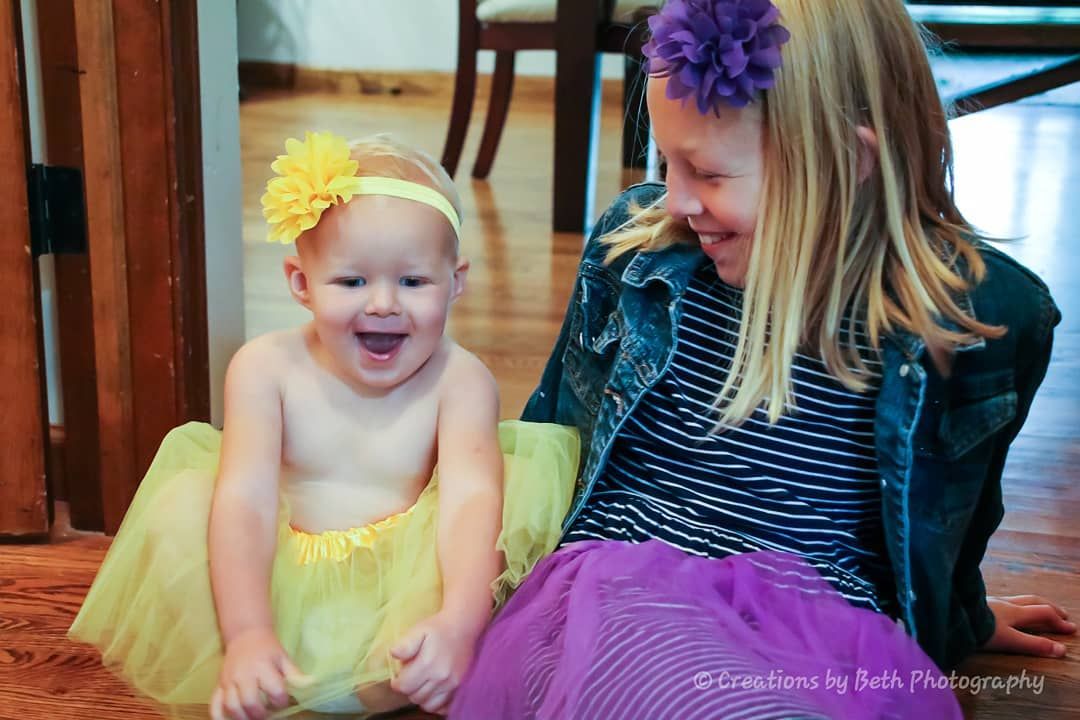 Two young girls smiling, wearing tutus and flower headbands. One is topless, seated on a wooden floor.
