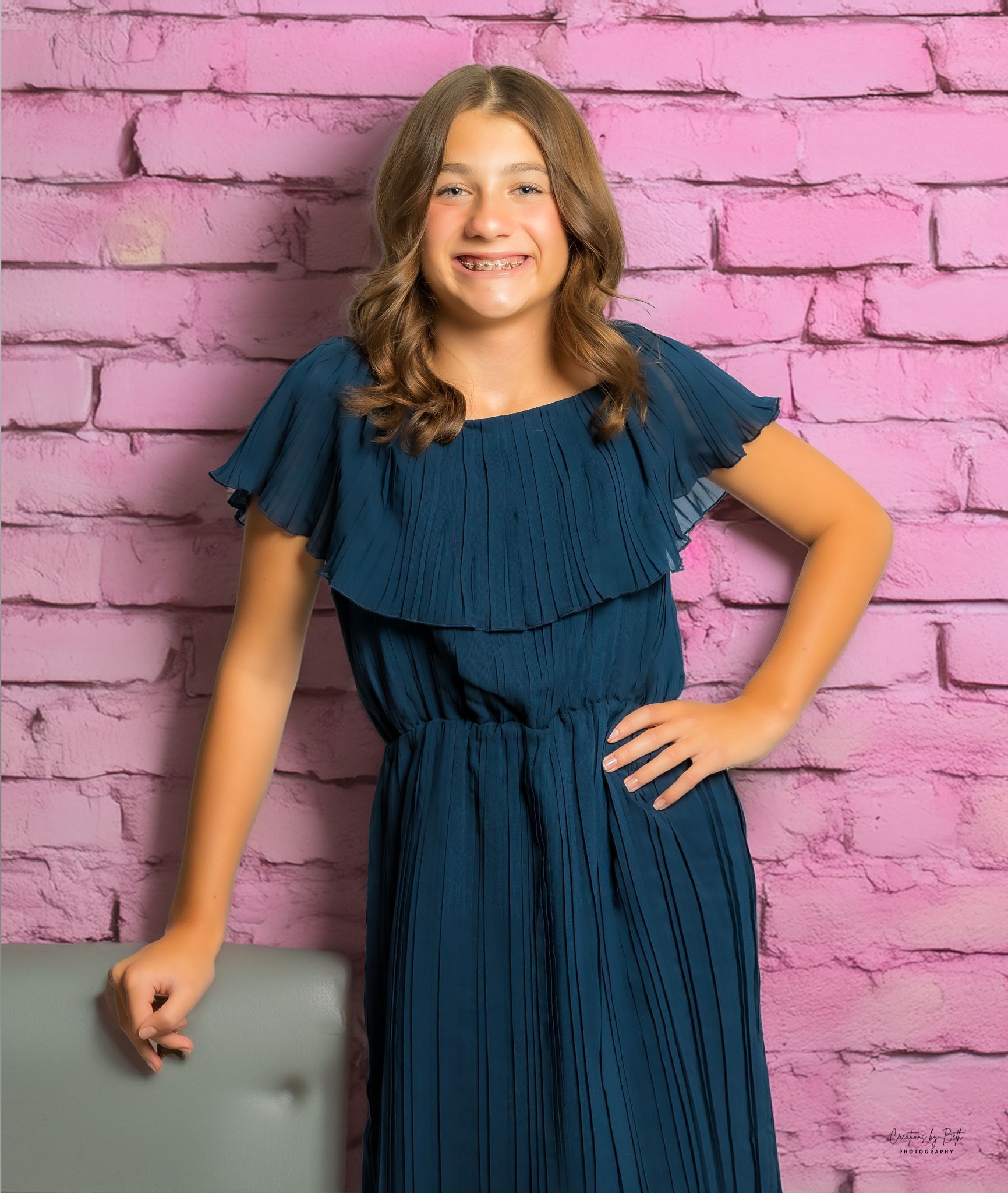 Young person in blue dress poses in front of a pink brick backdrop with hand on hip and a smile.
