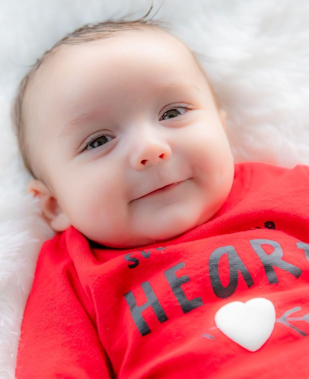 Baby in a red shirt with a white heart, smiling. Lying on a white, fluffy surface.