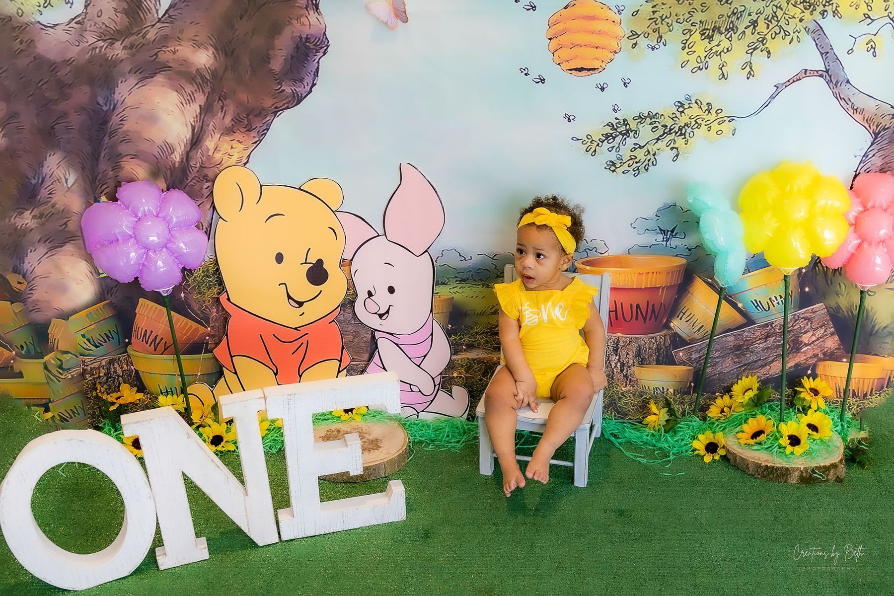 A child in yellow outfit sits in front of a Winnie the Pooh backdrop, surrounded by balloons and decor.