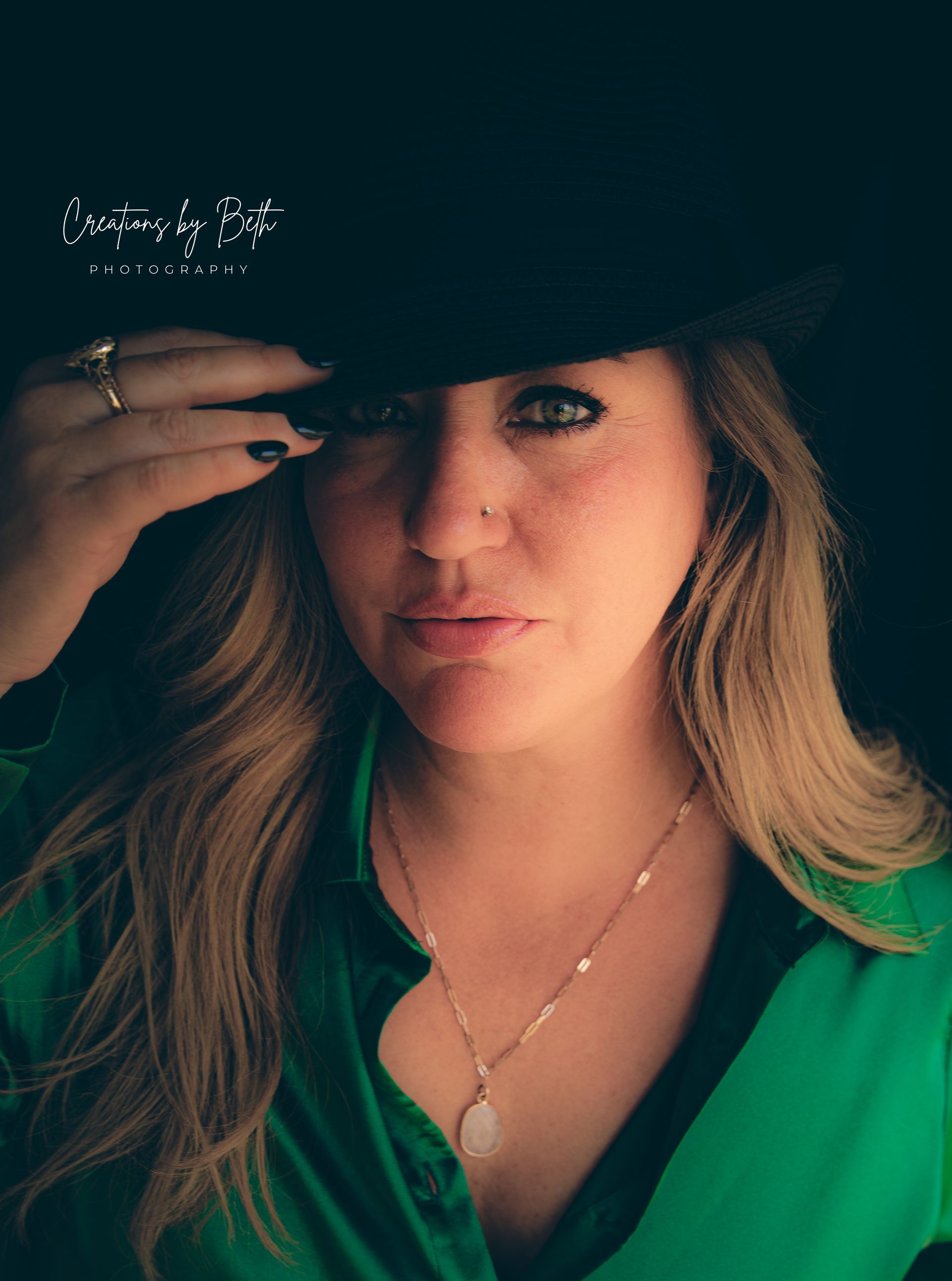 Woman in green shirt and black hat, looking at the camera, hand on the hat.