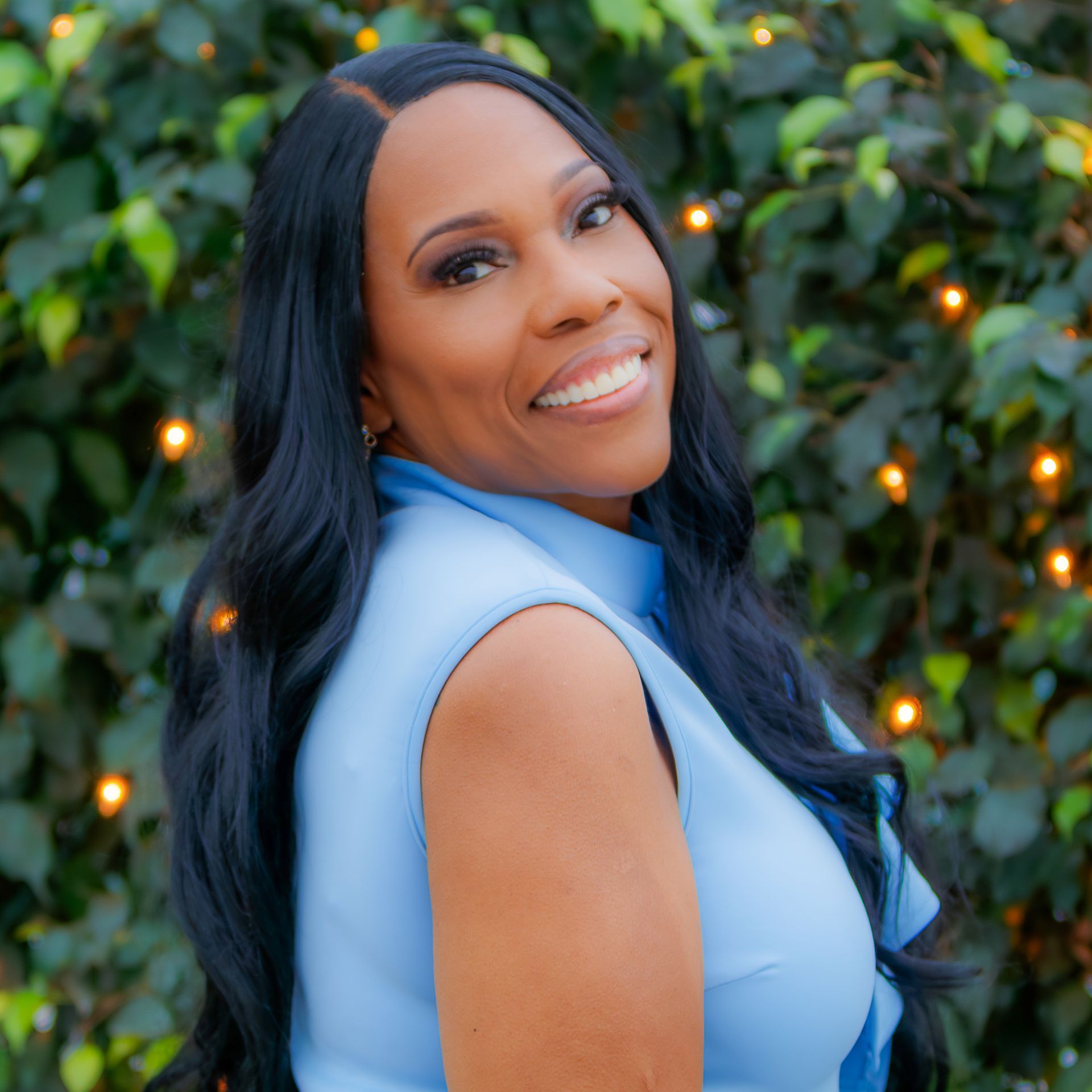 Woman in blue top smiles, dark wavy hair. Outdoor setting with greenery.