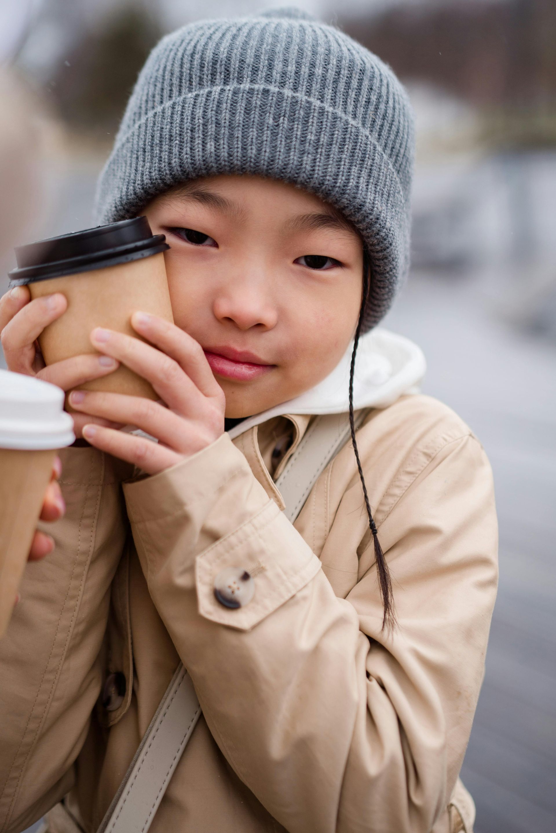 Person in a grey beanie holds two coffee cups, wearing a tan coat. Smiling, outdoors, blurred background.