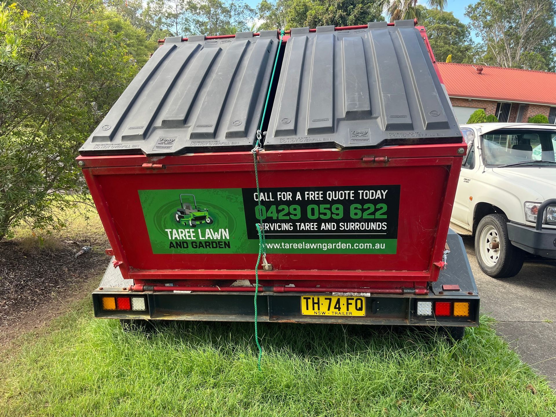 Red waste bin on trailer, with a green logo, parked on grass.