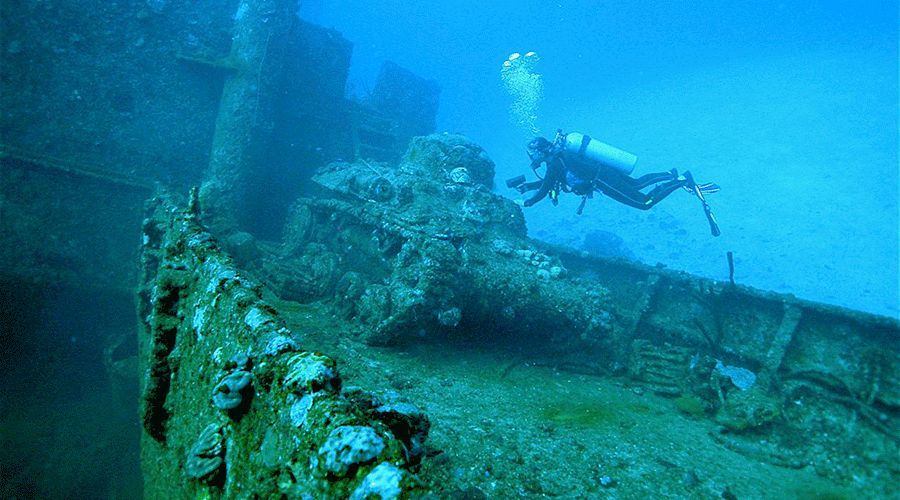 Scuba diver explores a weathered, submerged shipwreck in deep blue water.