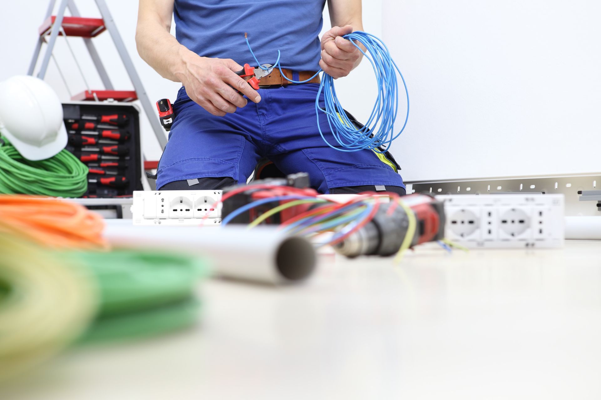 Electrician kneeling, cutting blue wires with pliers, surrounded by tools and cables.
