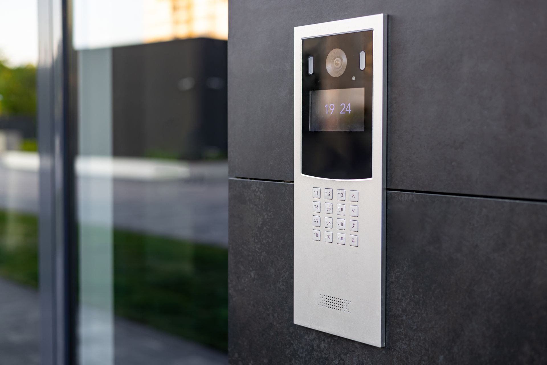 Silver intercom on a dark tiled wall; includes keypad, display, and camera.